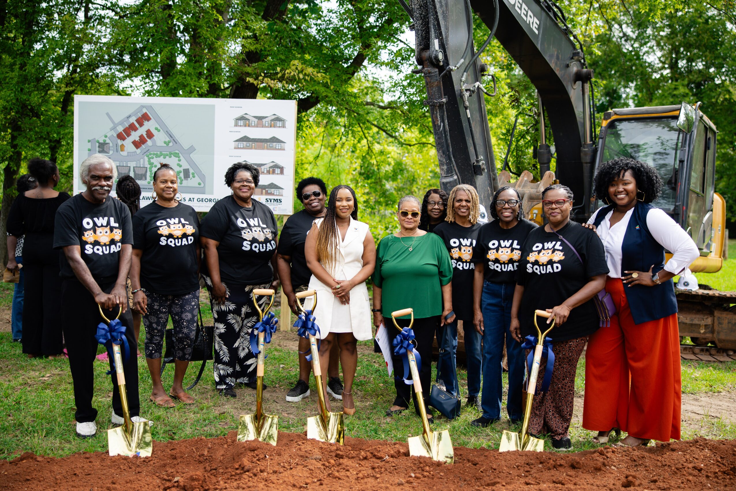 Group of people at a groundbreaking ceremony holding shovels, with trees and construction signs in the background.
