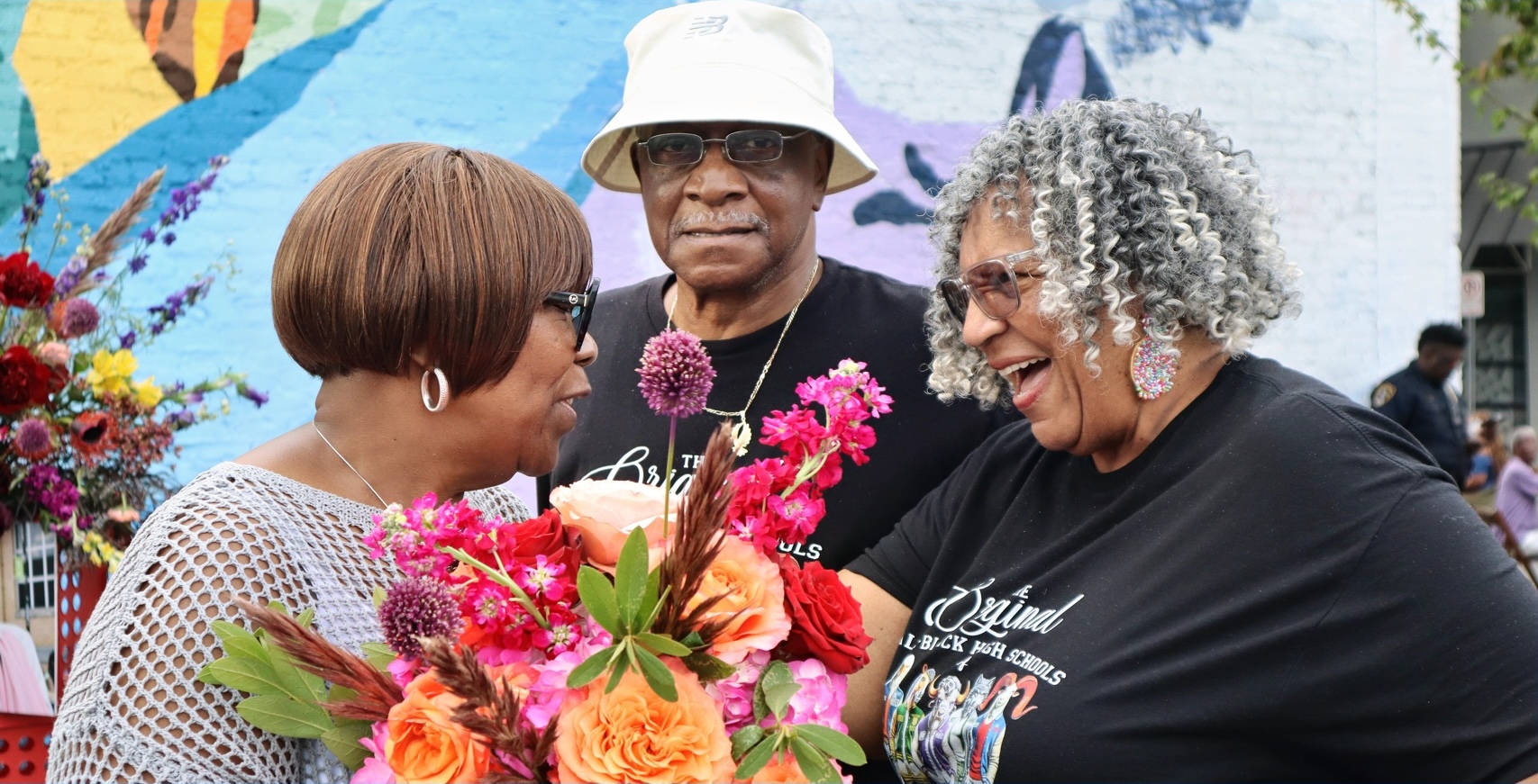 Three people smiling and exchanging flowers outdoors, with colorful background and flowers, one wearing glasses and a hat.