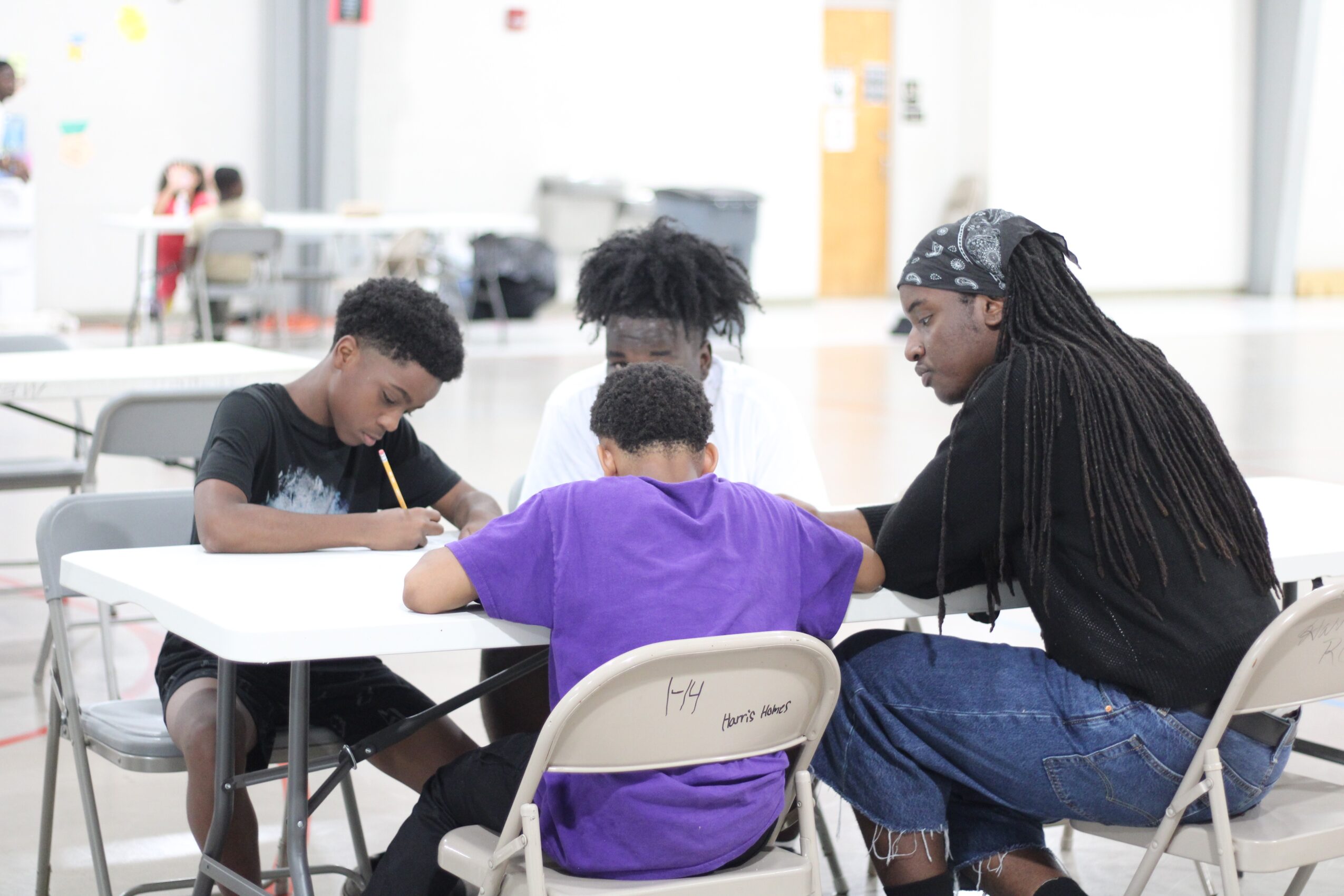Four people sitting at a table, drawing or writing, in a bright indoor space with large windows.