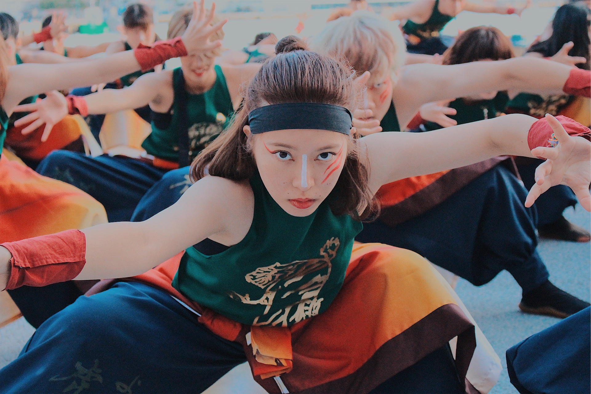 Group of people practicing yoga outdoors, with a woman in the foreground stretching forward, arms extended, wearing a headband and colorful clothing.