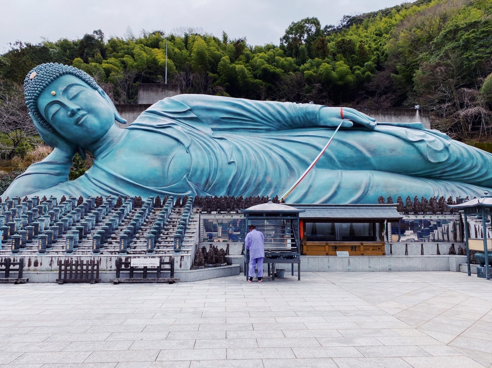 Large reclining blue Buddha statue outdoors with trees in background, person in white robes nearby, paved area in foreground.