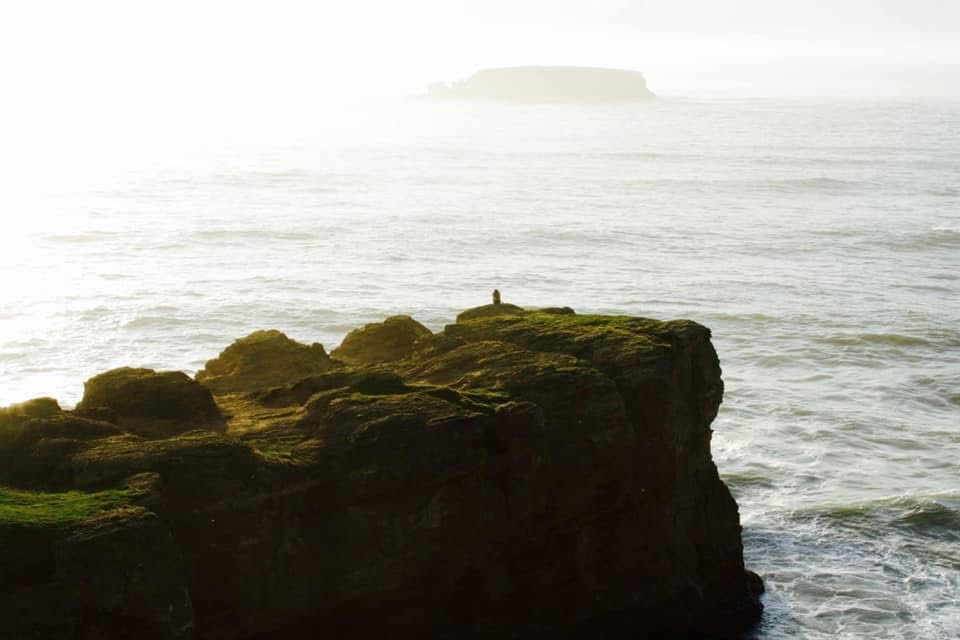 Cliff with a person standing on top overlooking the ocean, with a distant landmass in the background.