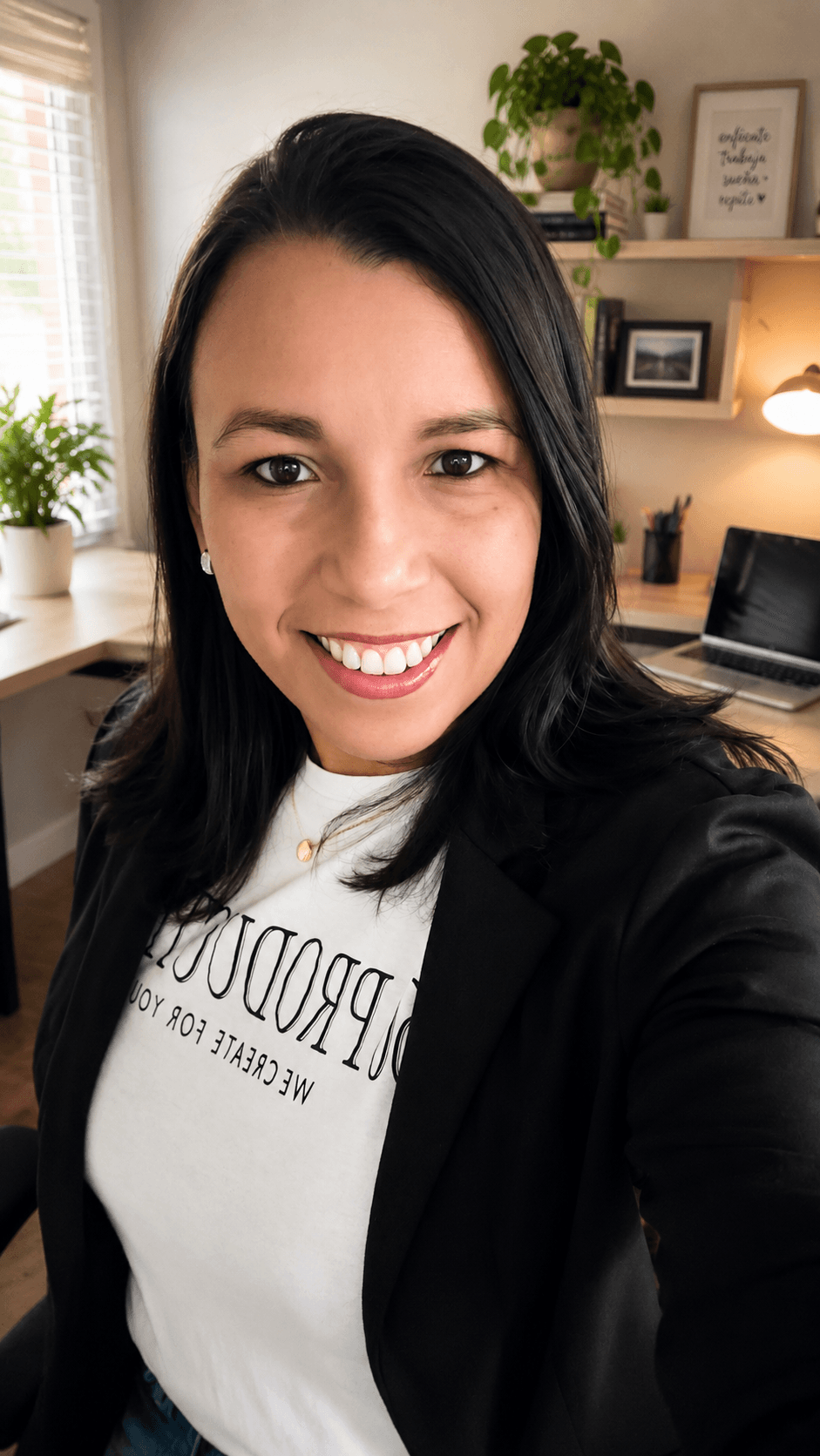 Smiling woman with dark hair in a home office, shelves with plants and framed pictures behind her.