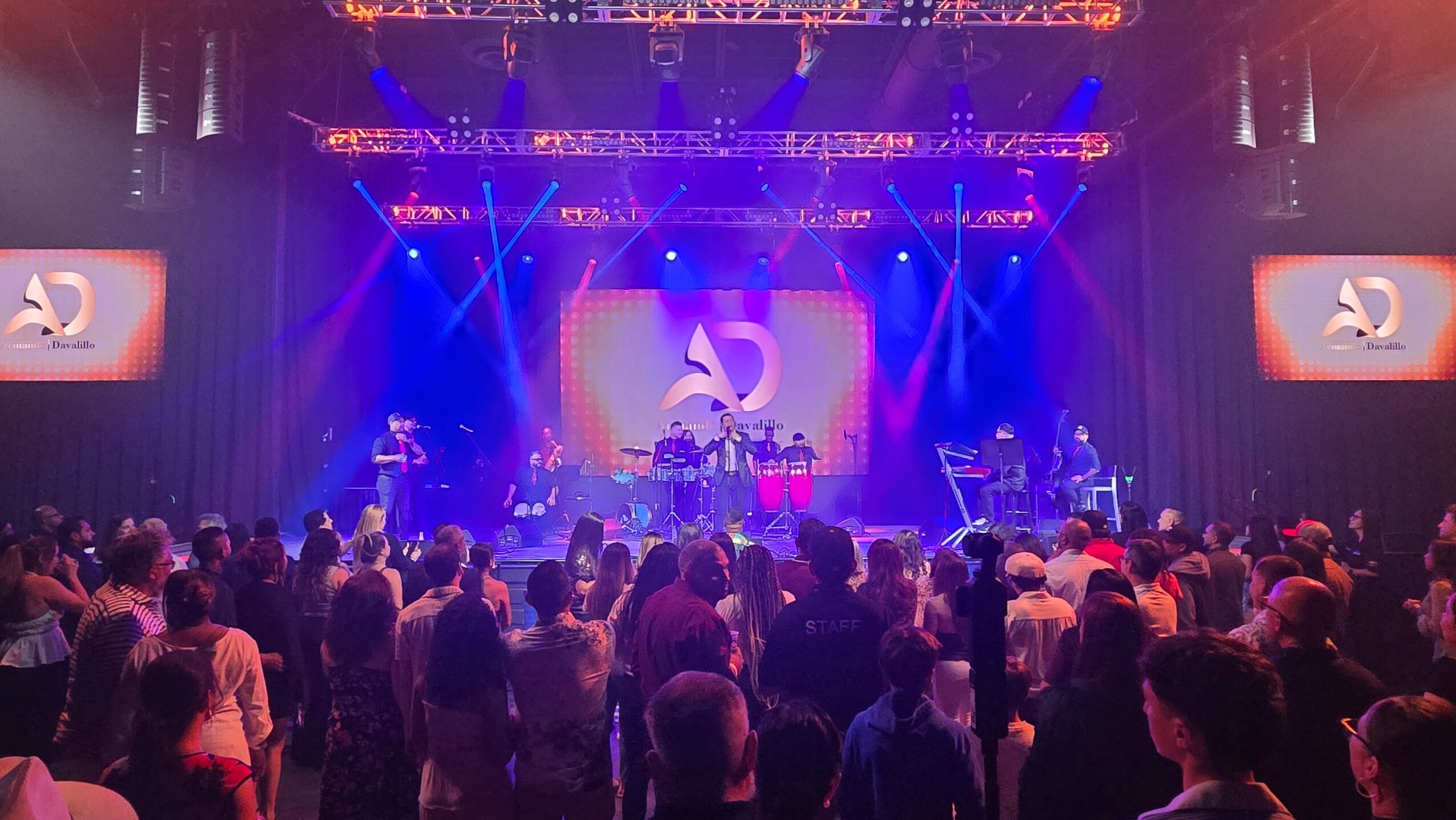 Crowd watching a stage with musicians and colorful lighting, screens displaying a logo, in a concert hall.