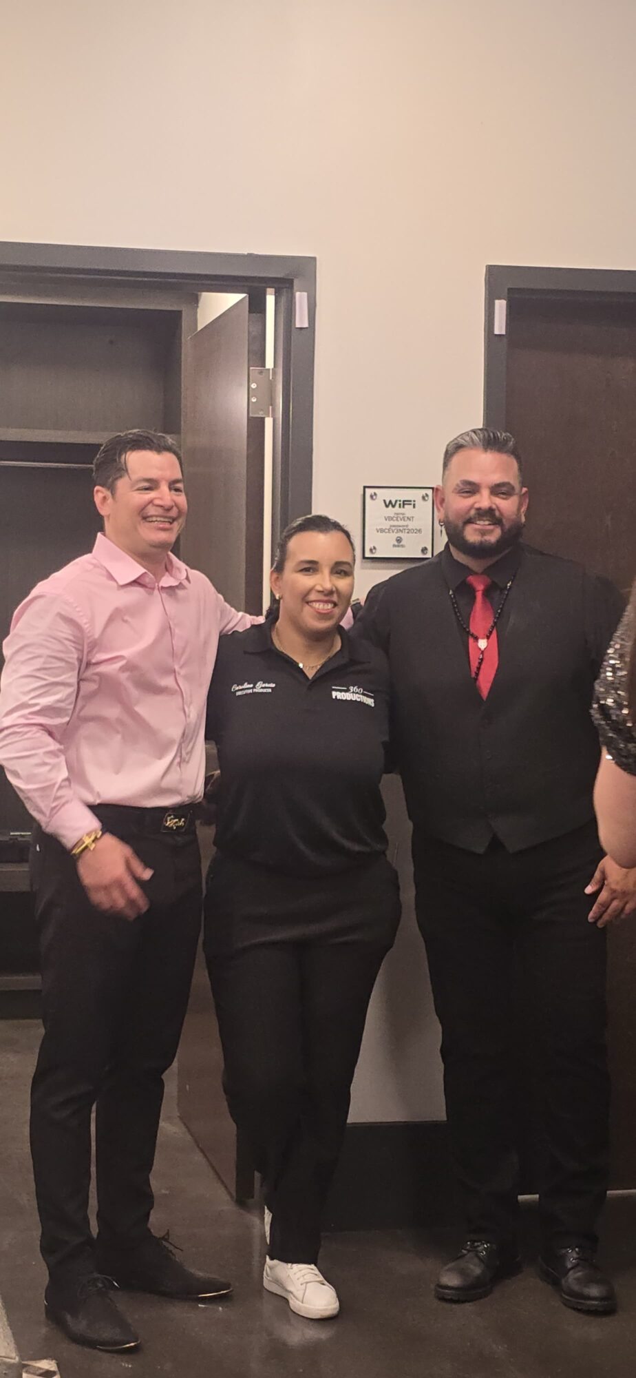 Three people standing indoors, smiling, with open doors behind them, one wearing a pink shirt, one in a black jacket, and one in a black suit with a red tie.
