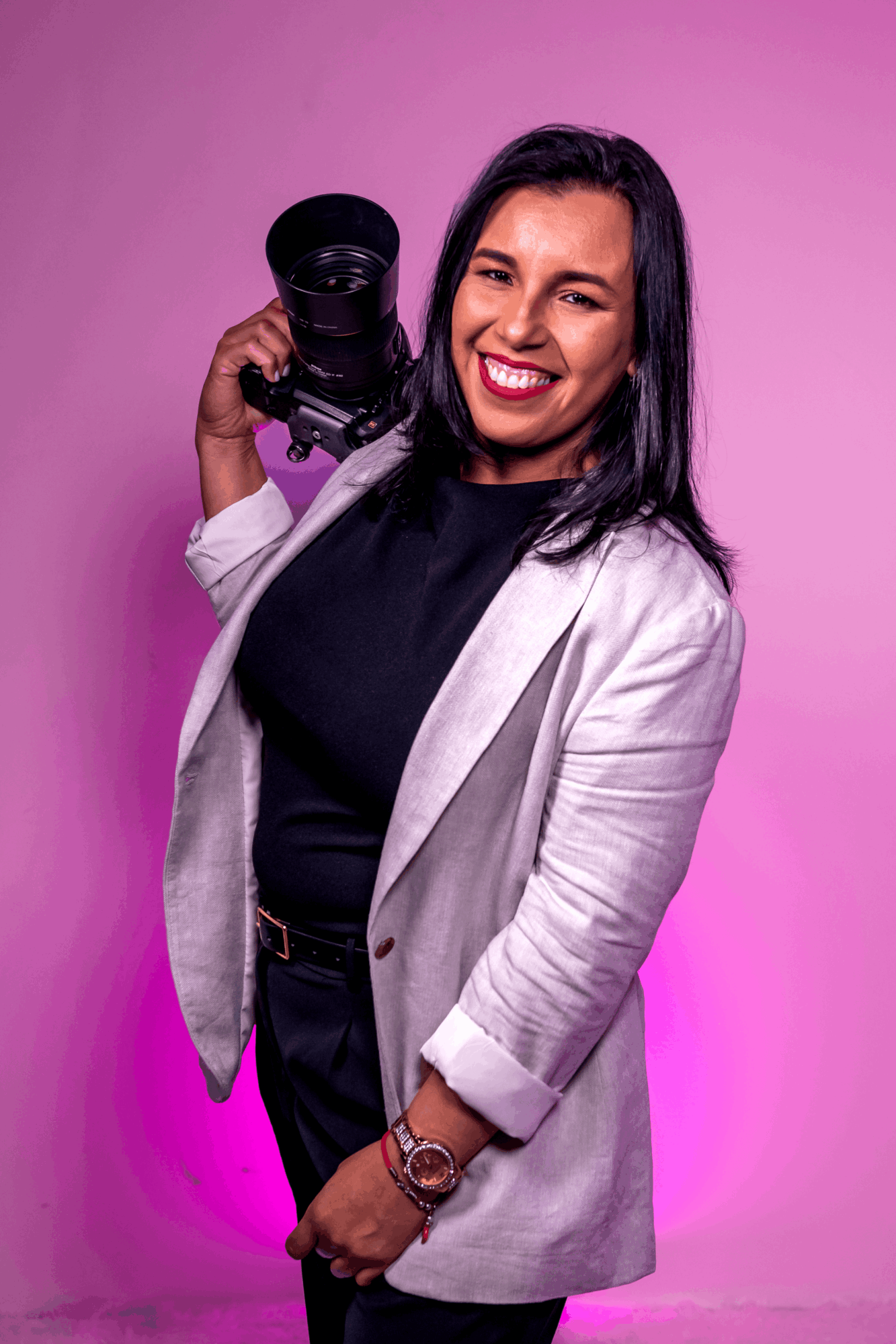 Woman with black hair smiling, holding a camera, wearing a light blazer and black top, against pink background.