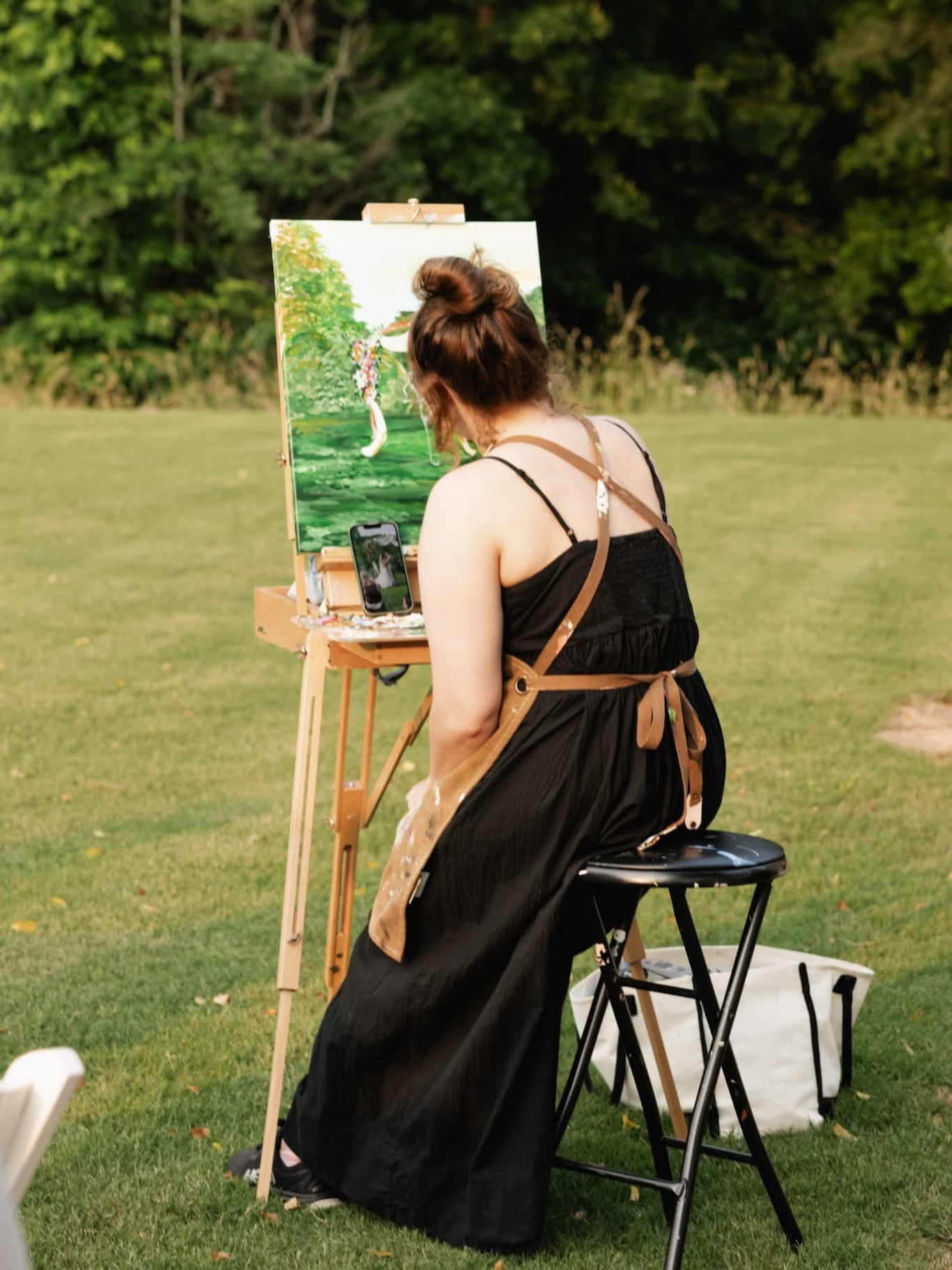Woman painting outdoors on an easel, sitting on a stool in a grassy area with trees in the background.