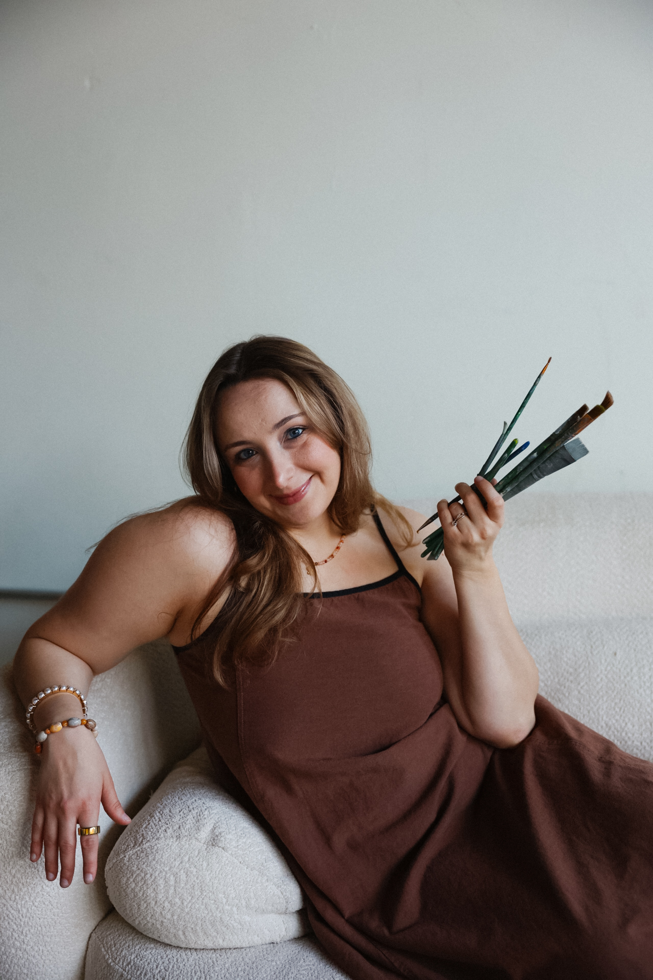 Woman with long hair in a brown dress sitting on a sofa holding paintbrushes and a palette, smiling.