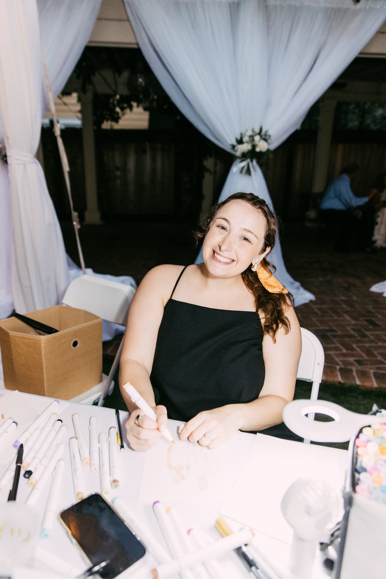 Young woman with long hair smiling, sitting at a table with makeup tools, outdoors with white curtains behind her.