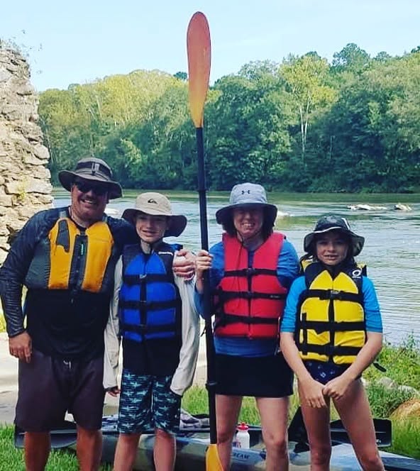 Four people in life jackets stand by a river with trees in the background, one holding a paddle. All are smiling.