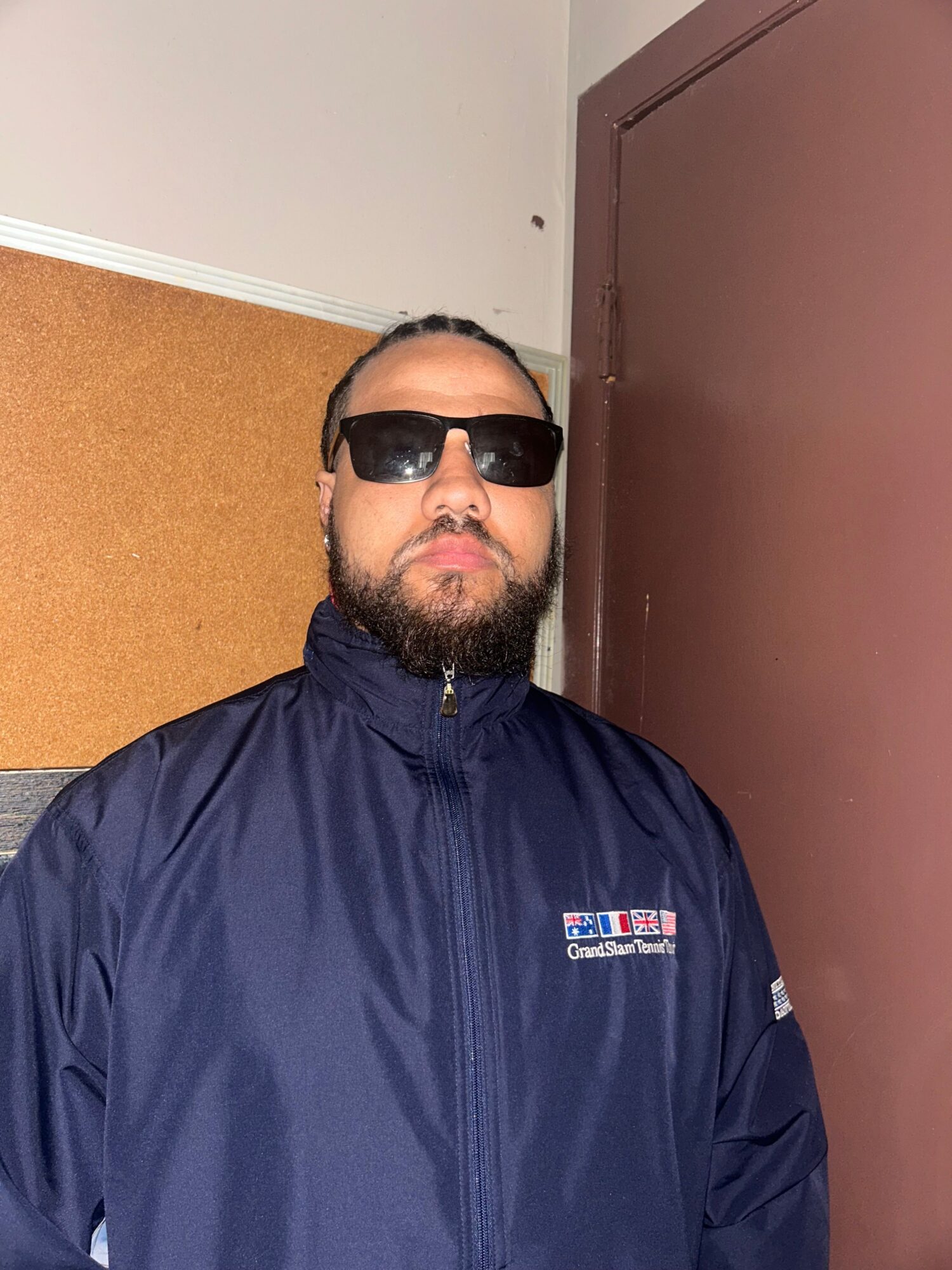 Man with beard wearing sunglasses and a dark jacket, standing indoors against a brown door and white wall.