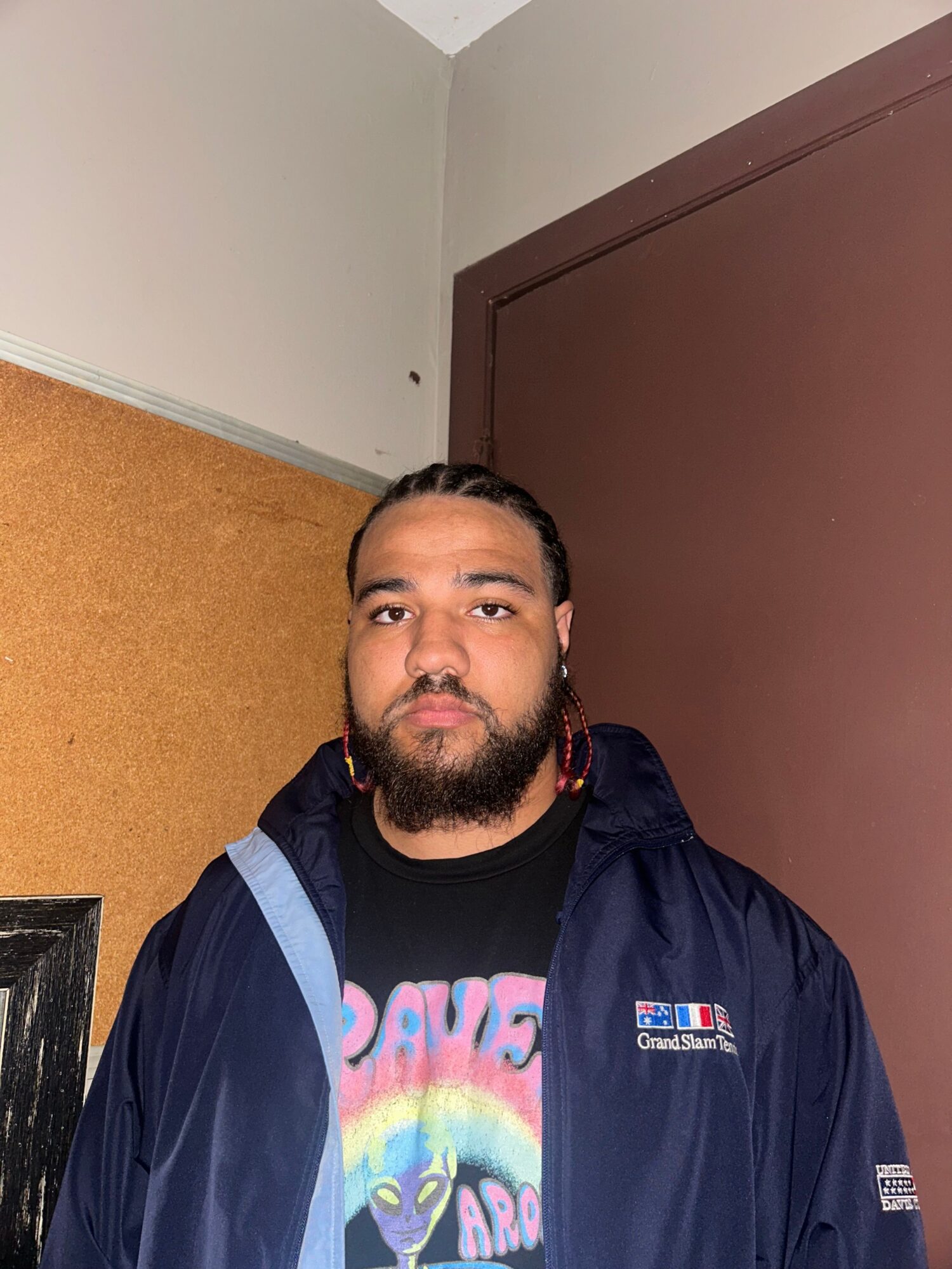 Man with beard and braided hair, wearing a dark jacket and colorful shirt, standing indoors near a brown door.