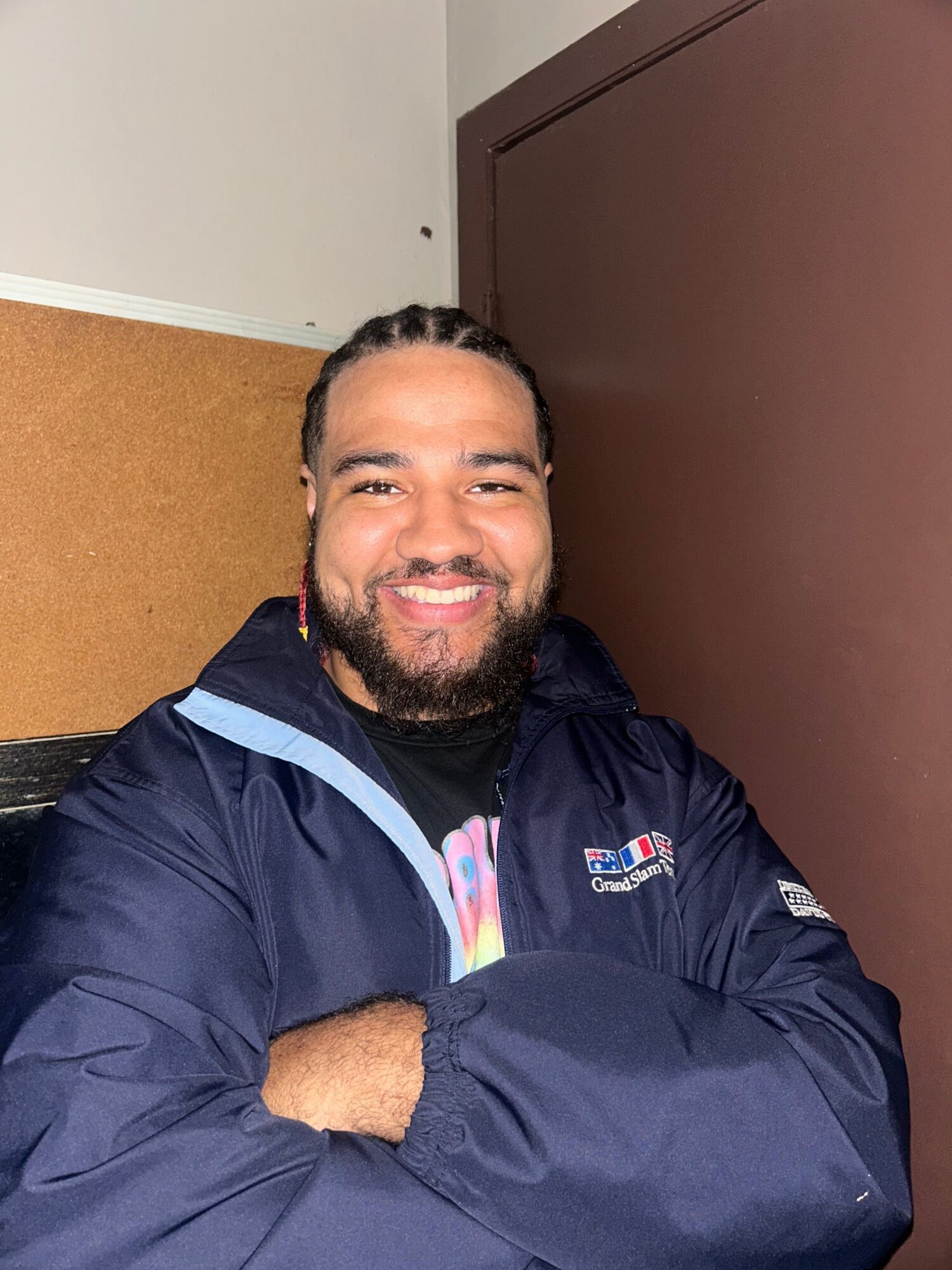 Person with a beard smiling, wearing a dark jacket, lying on a surface, with a brown door behind them.