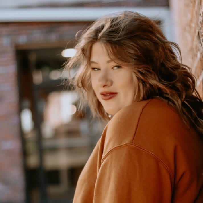 Woman with wavy hair wearing a brown top, looking to the side outdoors.