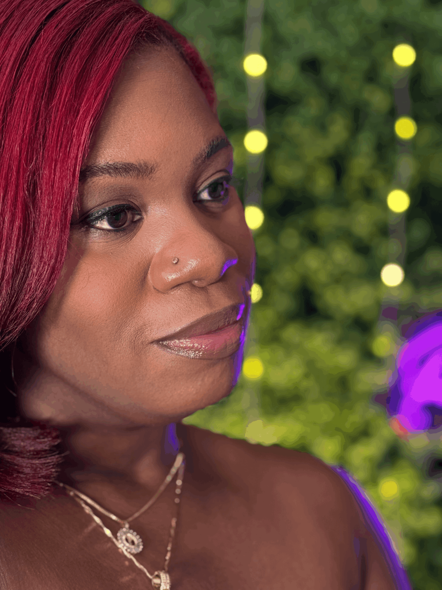 Close-up of a woman with red hair, wearing jewelry, outdoors with blurred green background and lights.