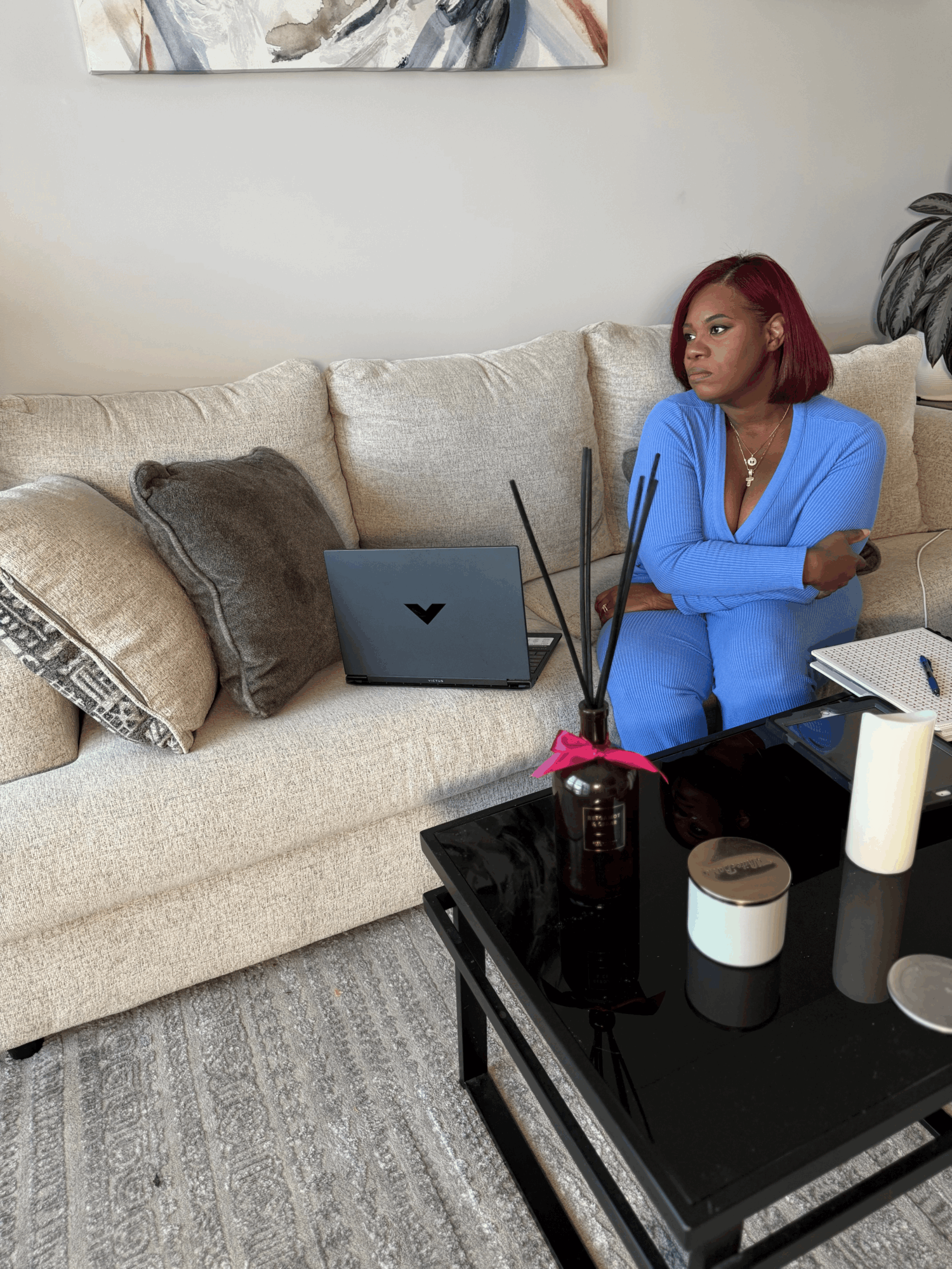 Woman with red hair sitting on a beige sofa, looking to the side, with a laptop and decorative pillows nearby.