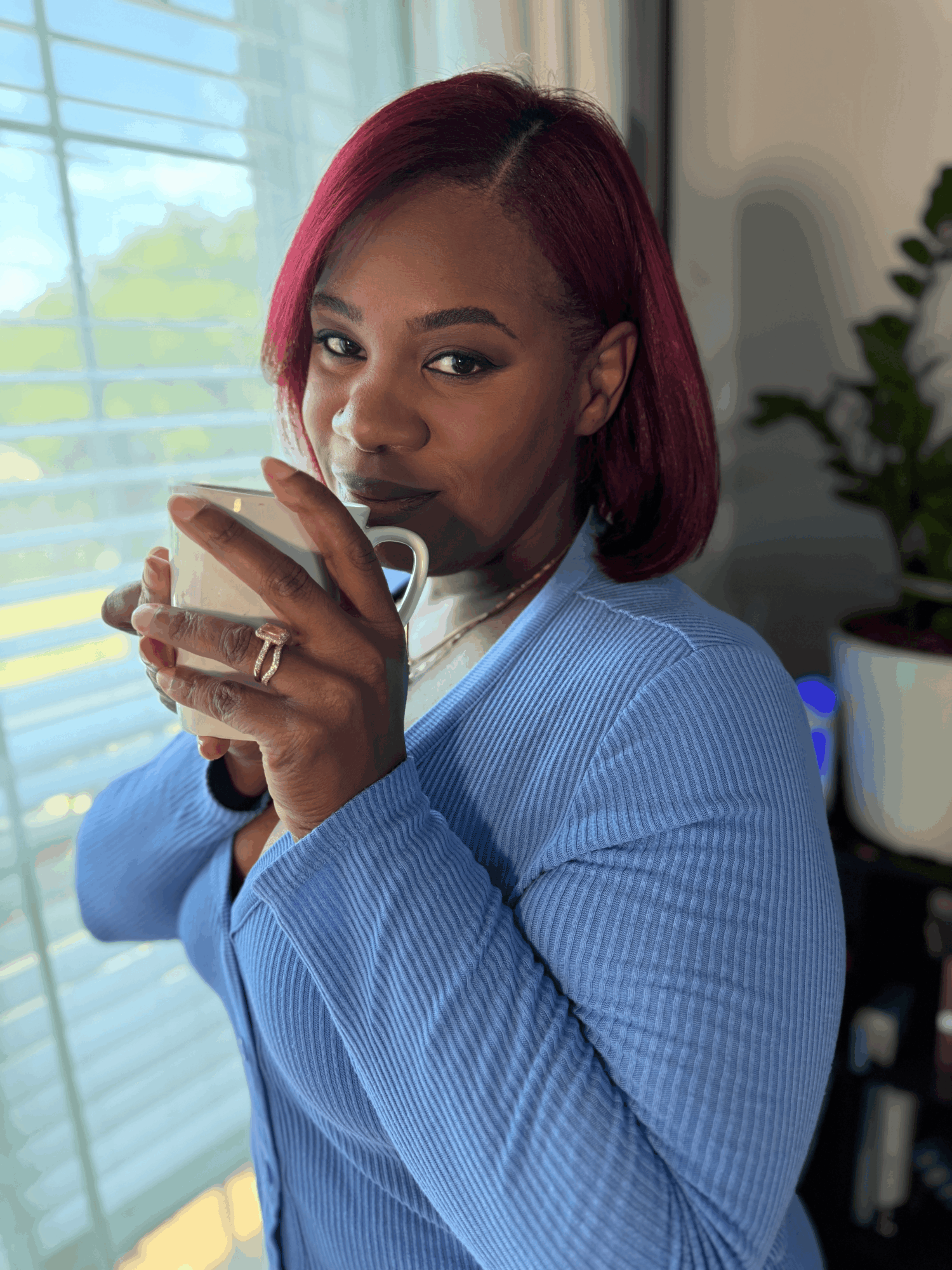 Woman with red hair holding a cup, standing near a window with blinds, in a room with a plant and bookshelf
