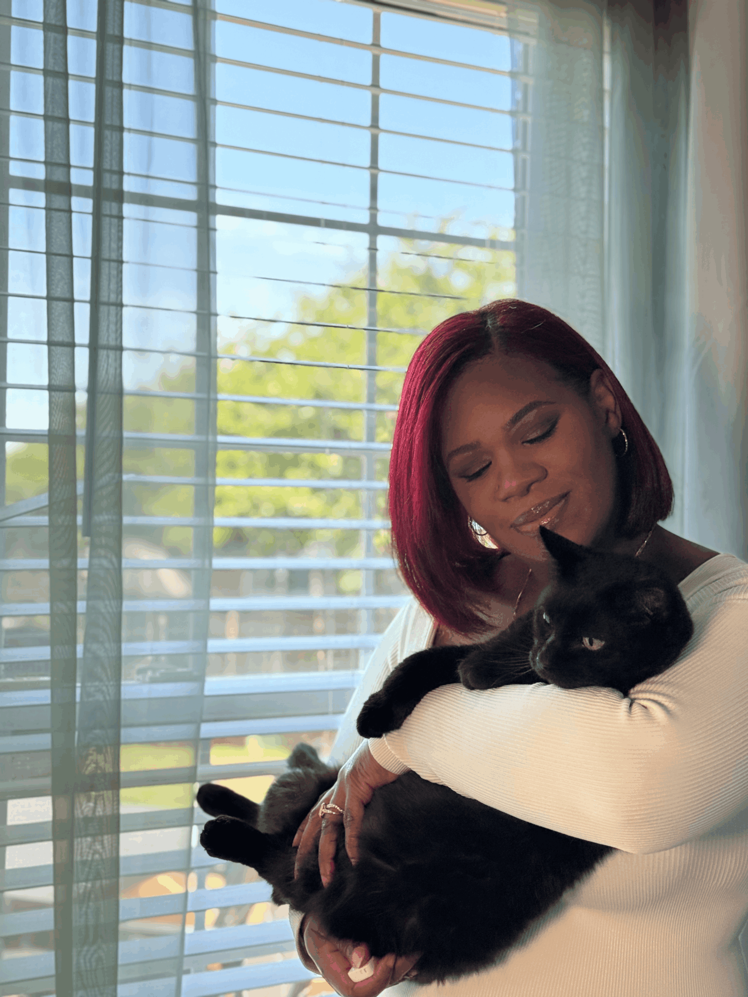 Woman with red hair holding a black and white cat near a window with blinds, smiling gently.