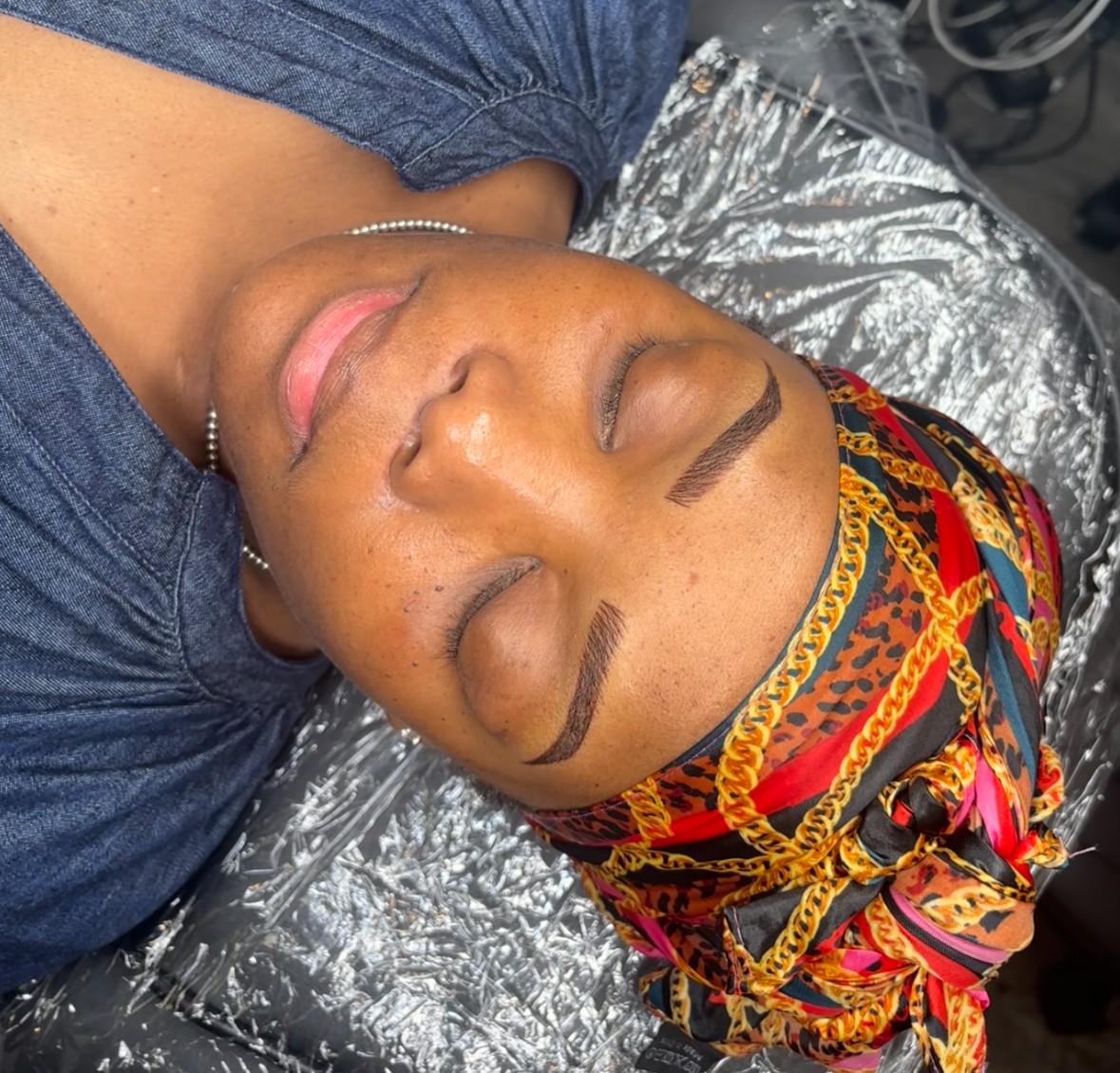 Woman with closed eyes, wearing colorful headscarf, lying down with makeup done, on a silver surface.