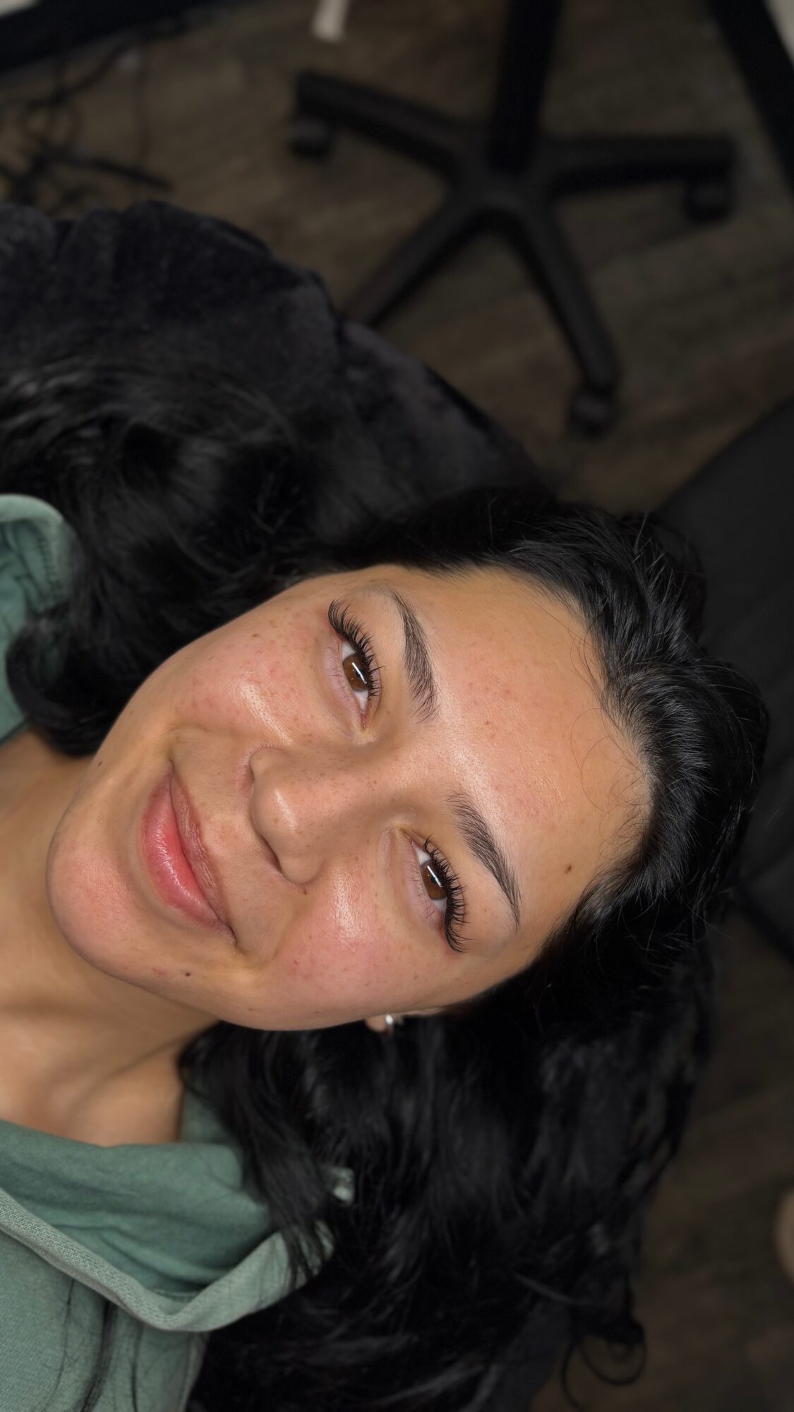 Woman with dark curly hair lying down, smiling, with a chair in the background.