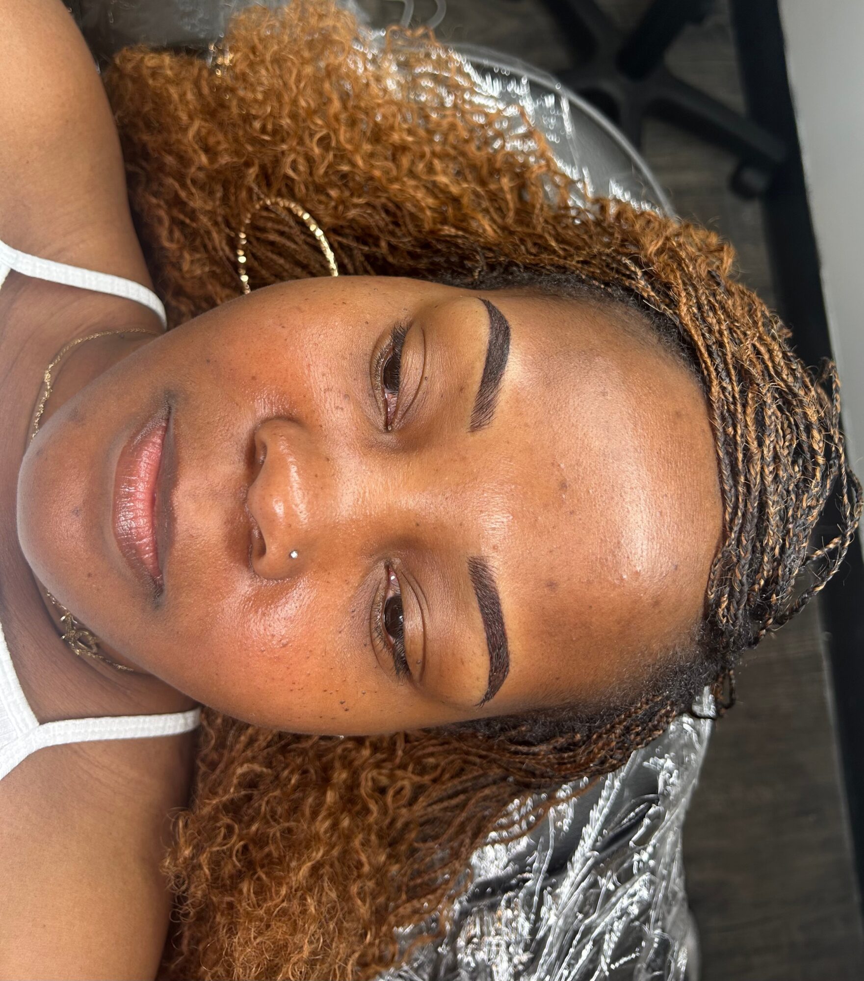 Close-up of a woman with curly hair and makeup, lying down with eyes closed, wearing a white top.