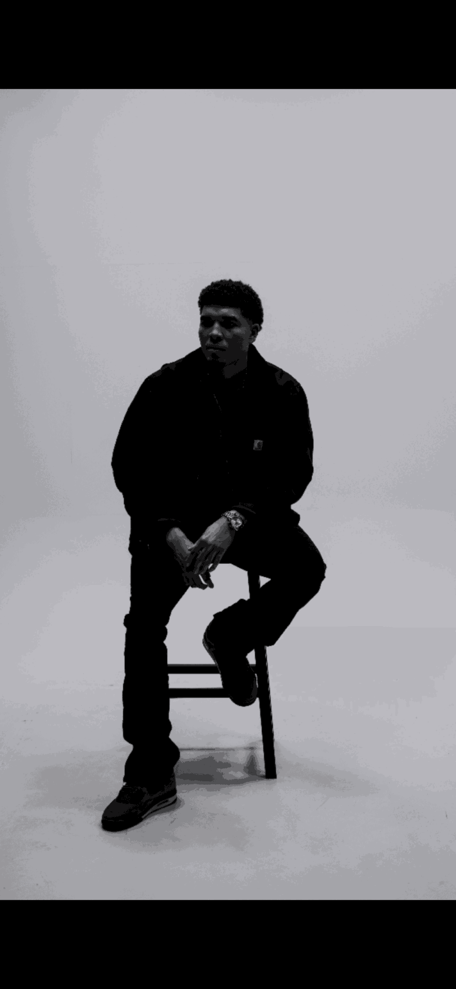 Person sitting on a chair in a plain room, facing forward, with hands clasped, in black clothing.