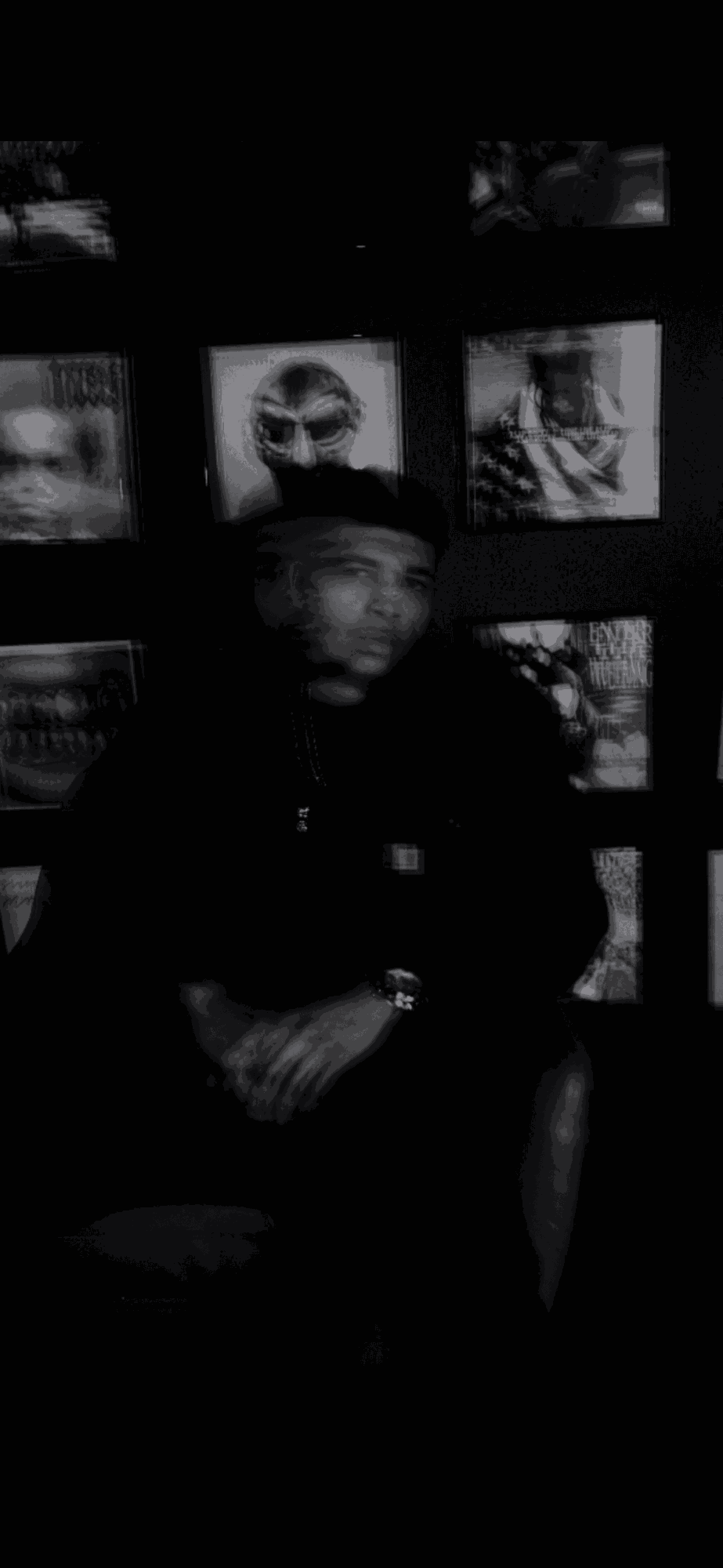 Person sitting in front of a wall with framed photographs, holding a drink, in a dimly lit environment.
