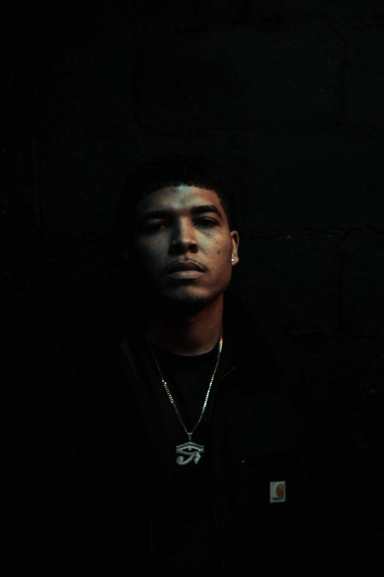 A young man with short hair and a serious expression, wearing a necklace, in a dark setting.
