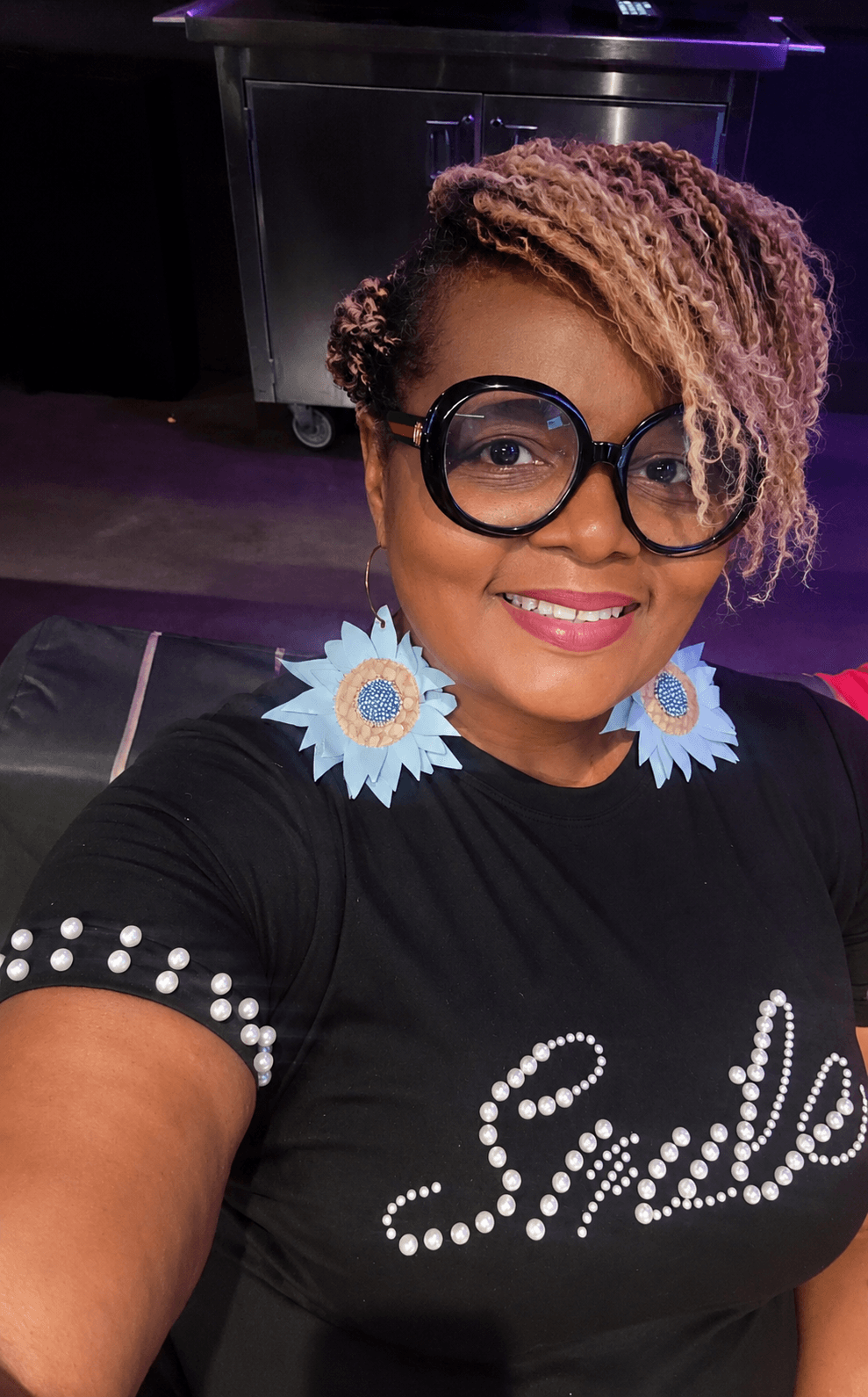 Smiling woman with short, curly hair, wearing large glasses and blue flower earrings, taking a selfie indoors.