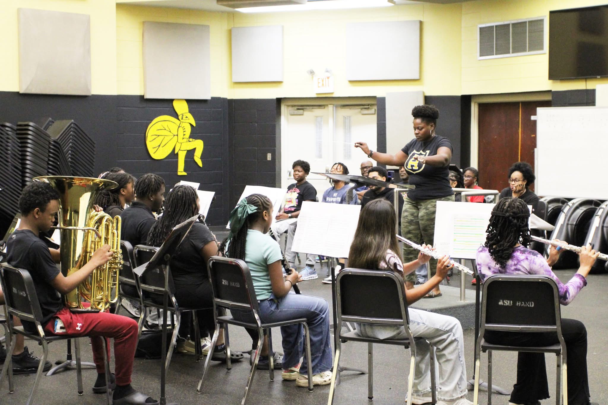 Music class with students playing wind instruments and a teacher conducting in a classroom with yellow and black walls.
