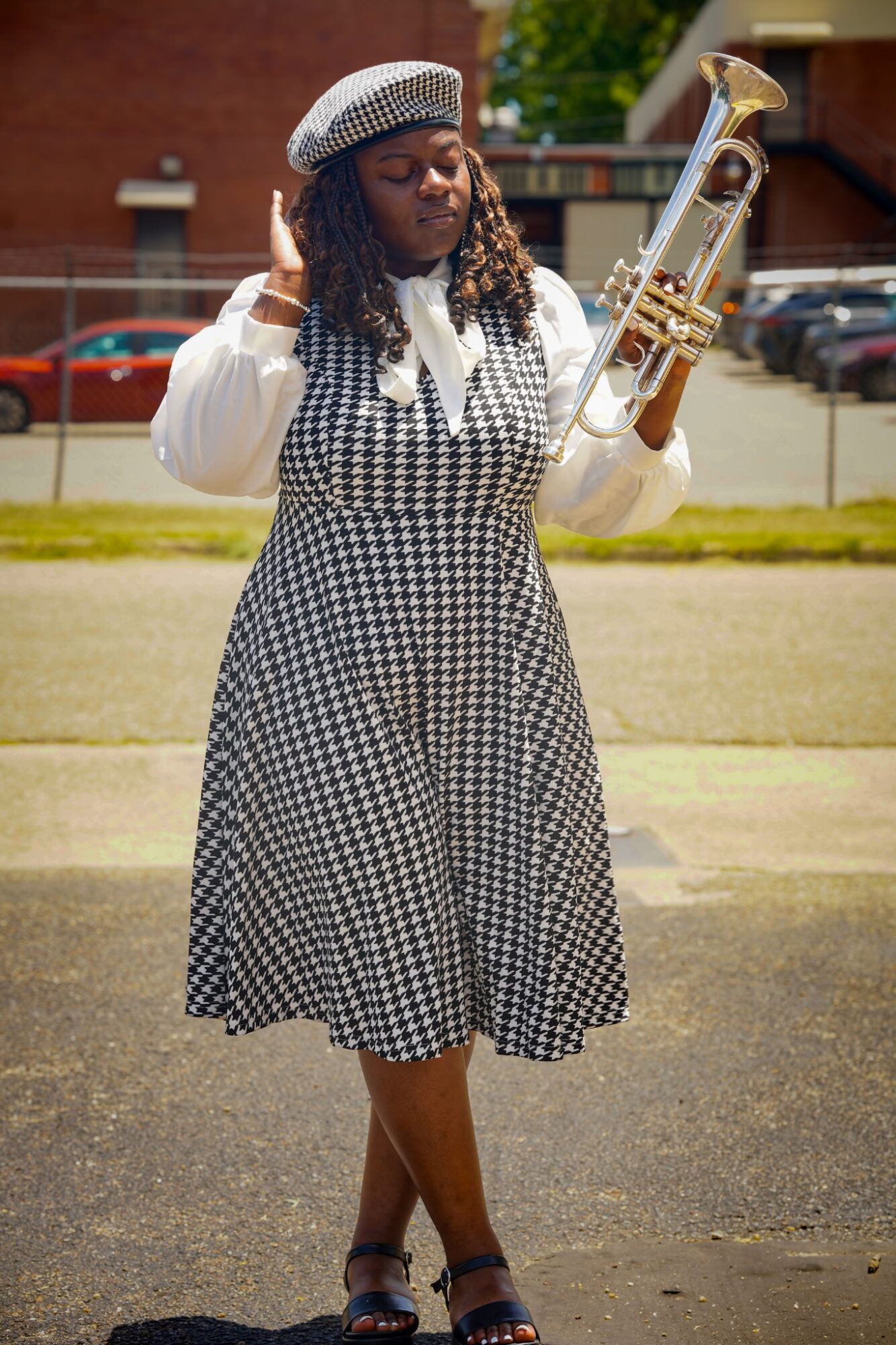 Woman in checkered dress and beret holding a saxophone outdoors, standing on grass with cars and buildings in background.