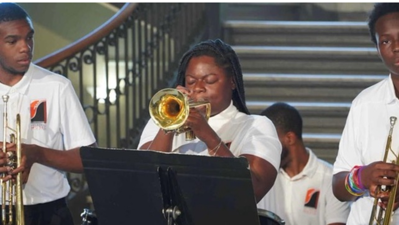 Group of young musicians playing brass instruments, standing on stairs with a railing in the background.