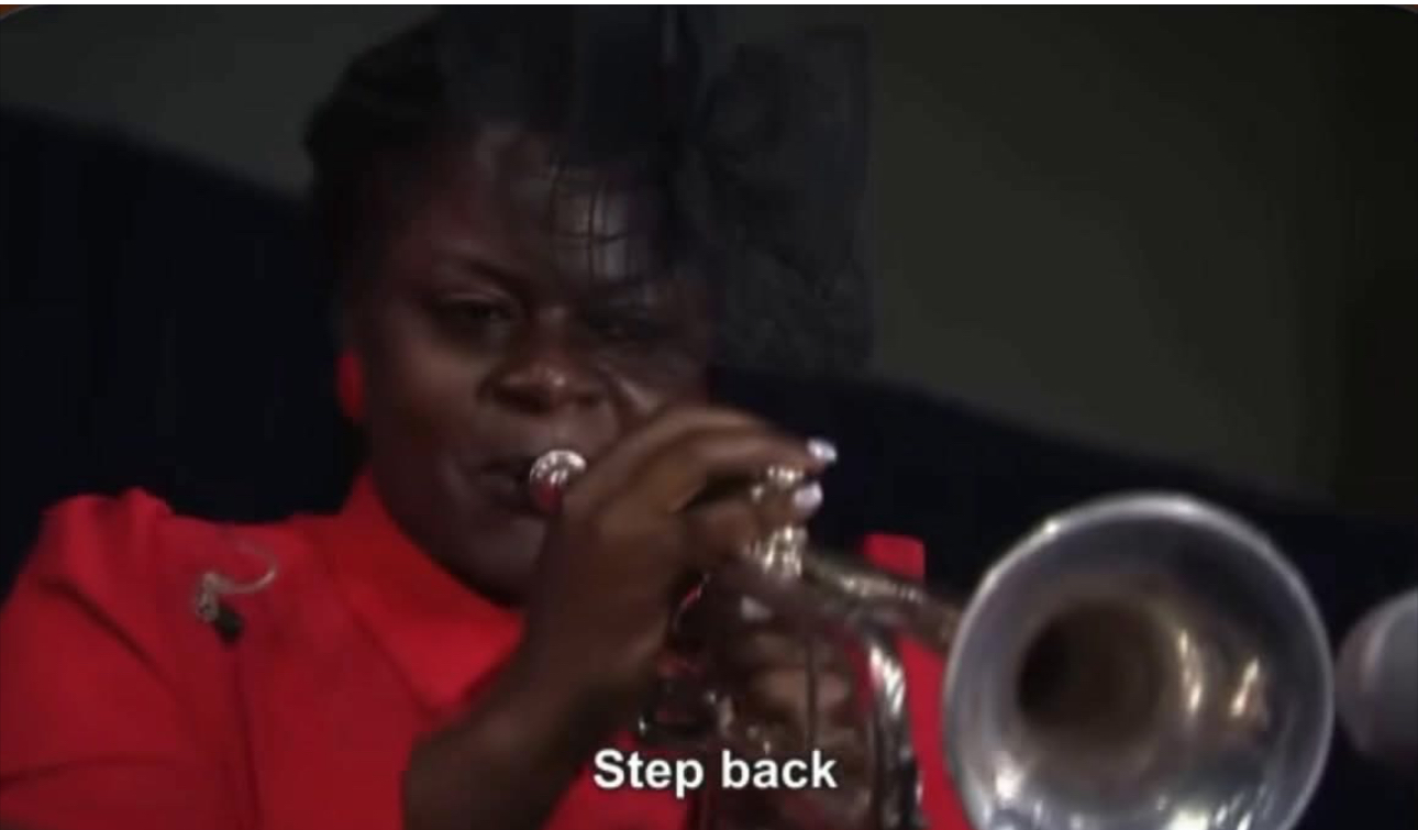 Person playing trumpet, wearing red clothing, with a focused expression, indoors, dark background.