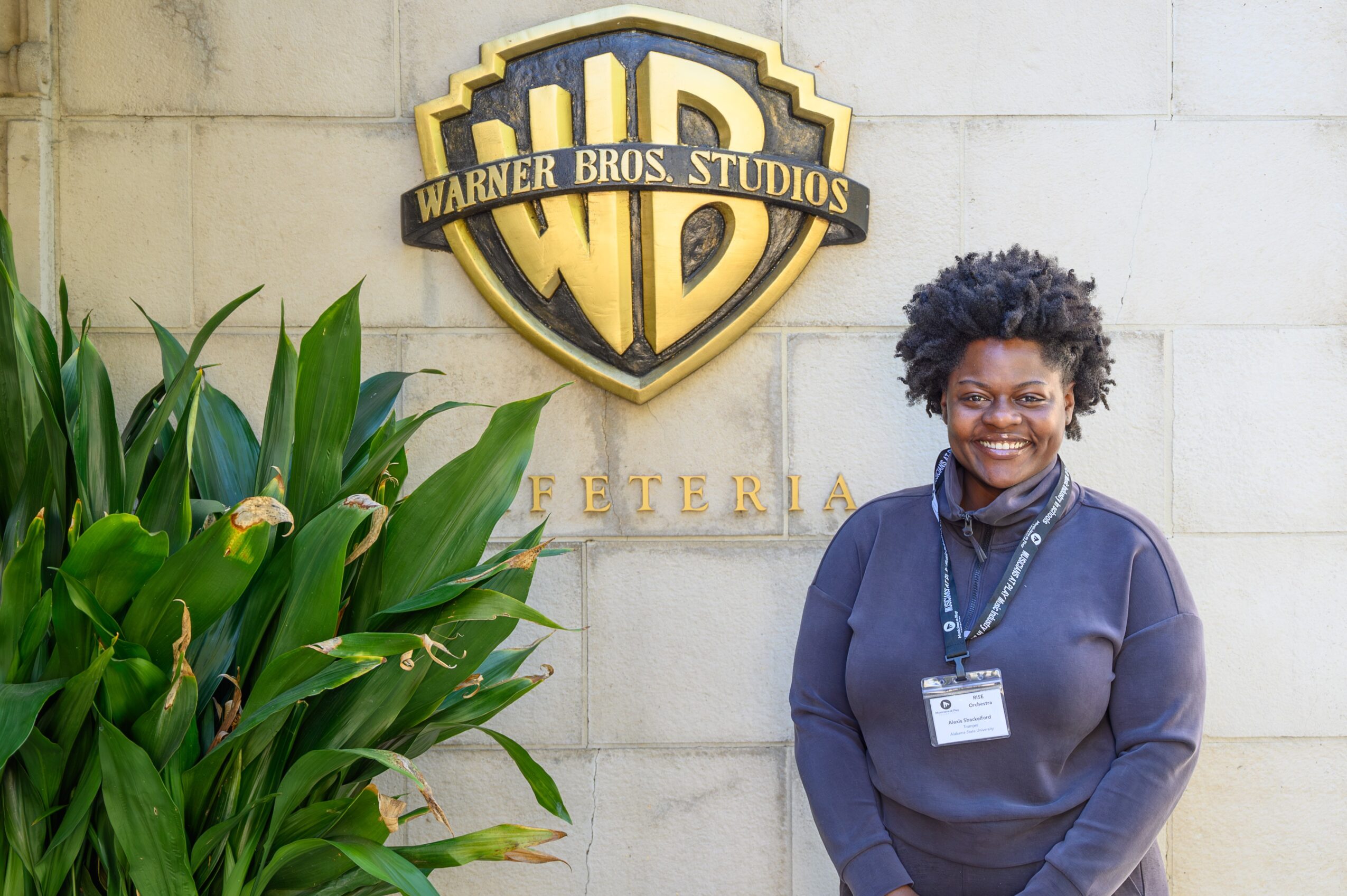 Woman with curly hair smiling standing in front of Warner Bros. Studios sign and greenery, wearing a dark sweater and lanyard.