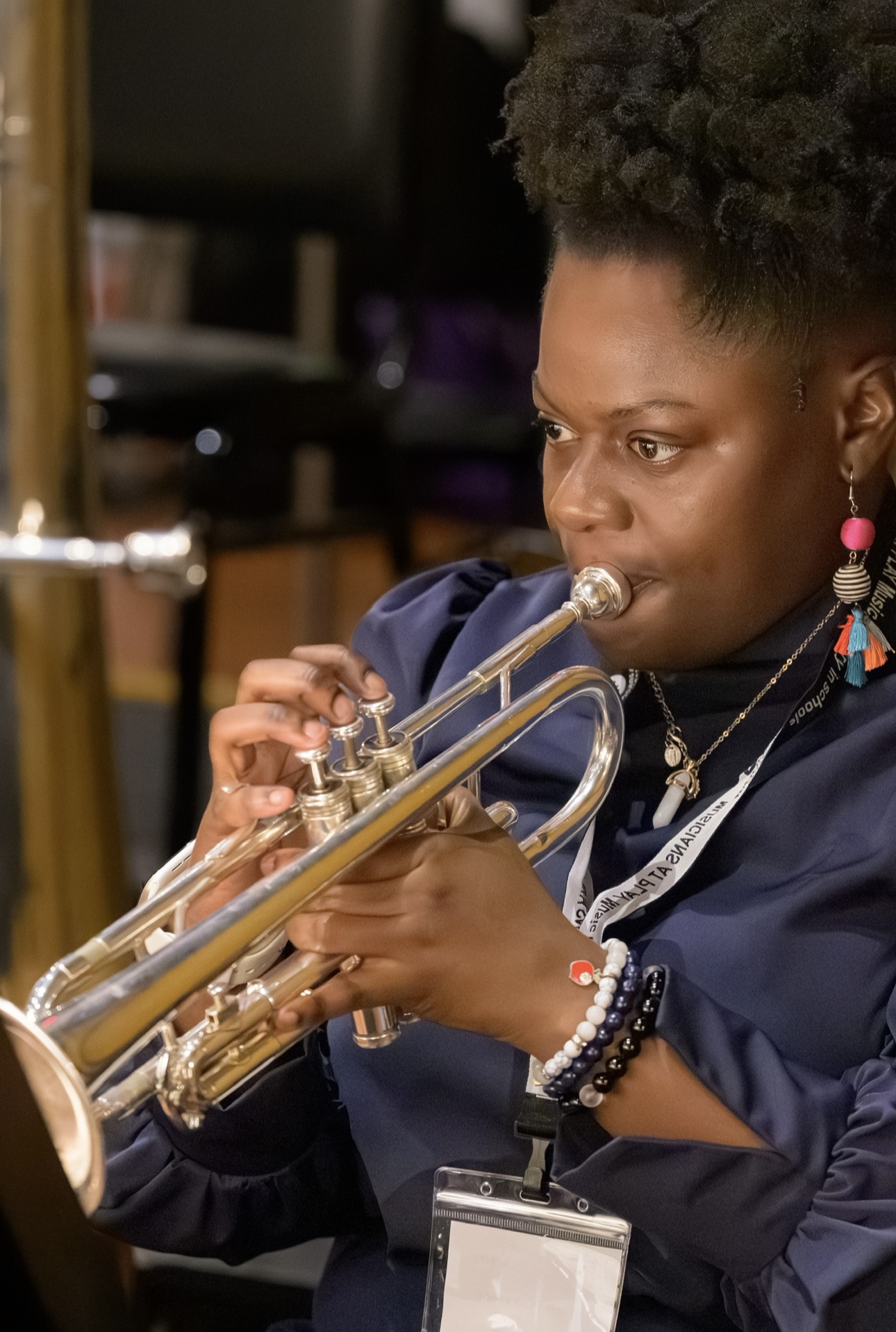 Woman playing trumpet, focused, indoors, wearing earrings and bracelets, with a blurred background.