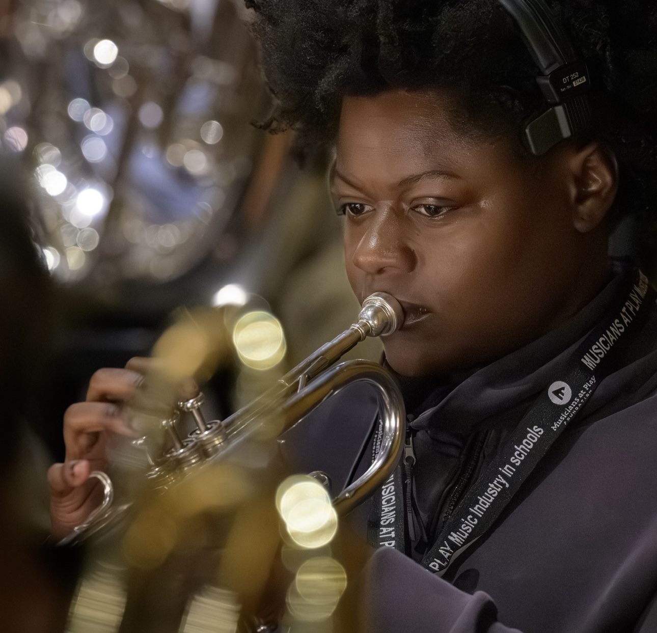 Person playing trumpet, focused expression, blurred background with lights, wearing headphones and dark clothing.