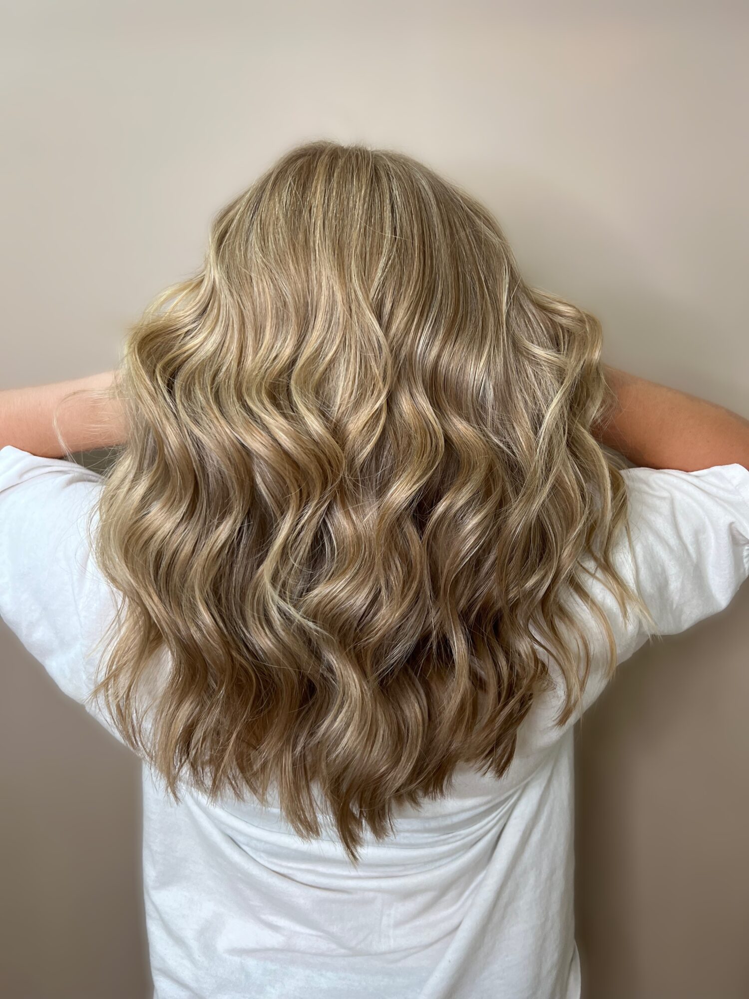 Back of a person with long, wavy blonde hair, wearing a white shirt, standing against a plain wall.