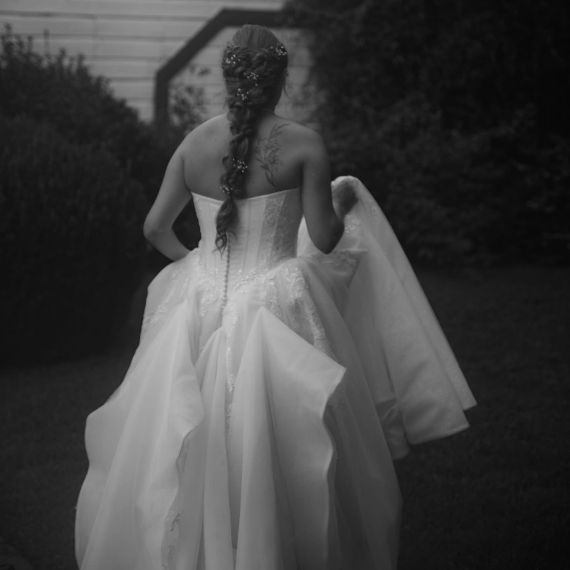 Back view of a woman in a wedding dress holding the skirt, with a braid hairstyle, outdoors in a garden.