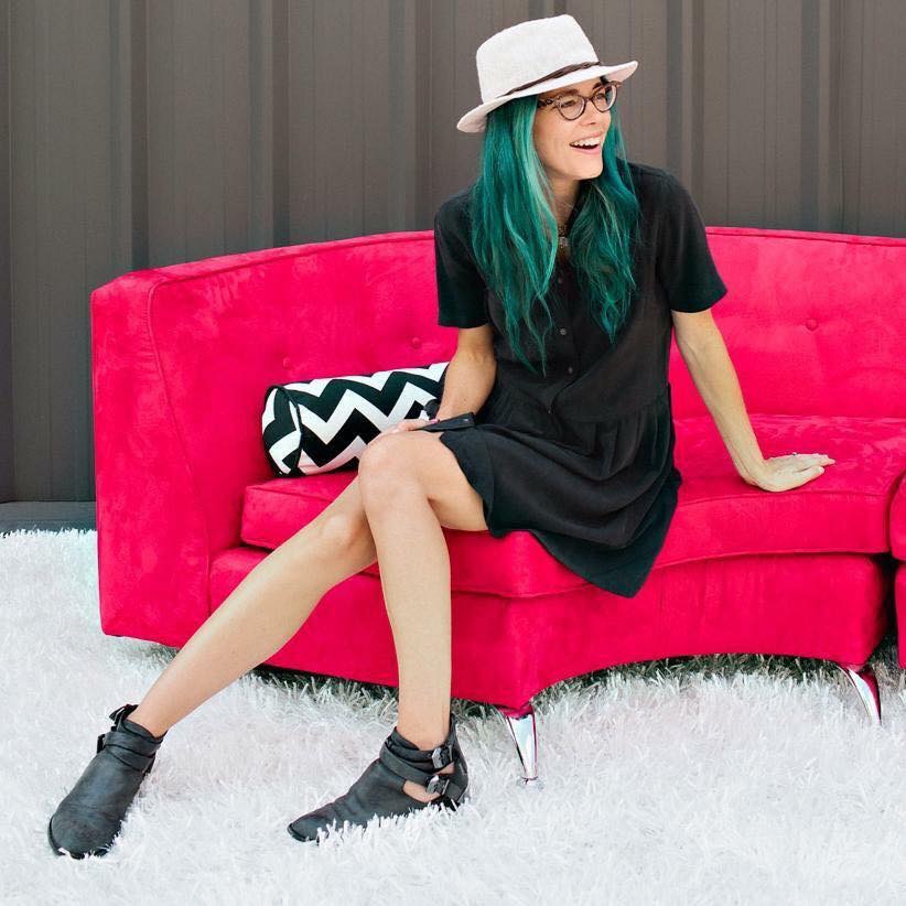 Woman with green hair, glasses, hat, black dress, sitting on a pink sofa, smiling, with a black and white pillow, white rug.
