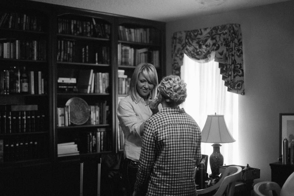 Two women talking in a room with a bookshelf, window with curtains, and a table lamp.