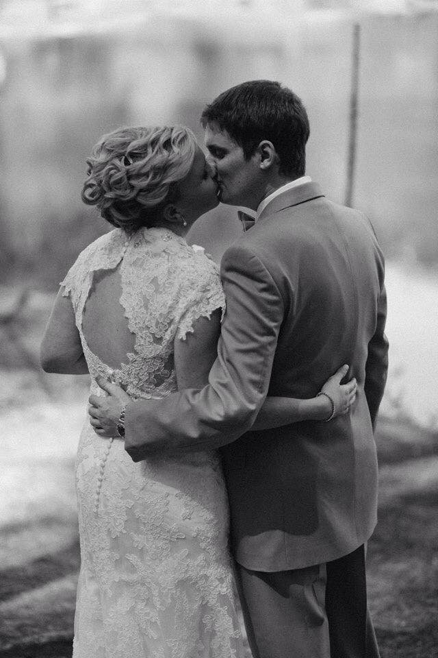 A couple in wedding attire kissing outdoors, the woman in a lace dress and the man in a suit, embracing each other.