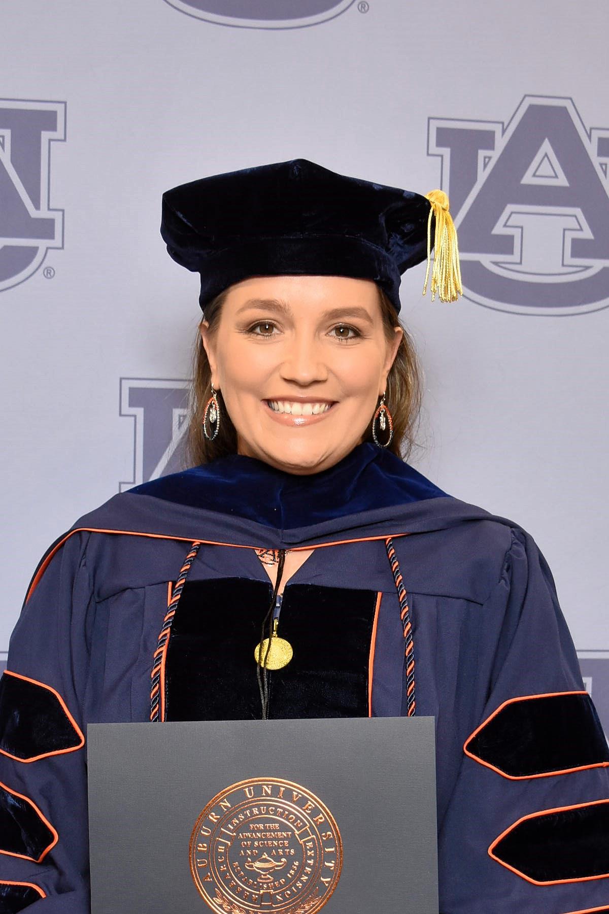 Woman in graduation cap and gown smiling, holding diploma, background with university logo, wearing earrings, with a yellow tassel.