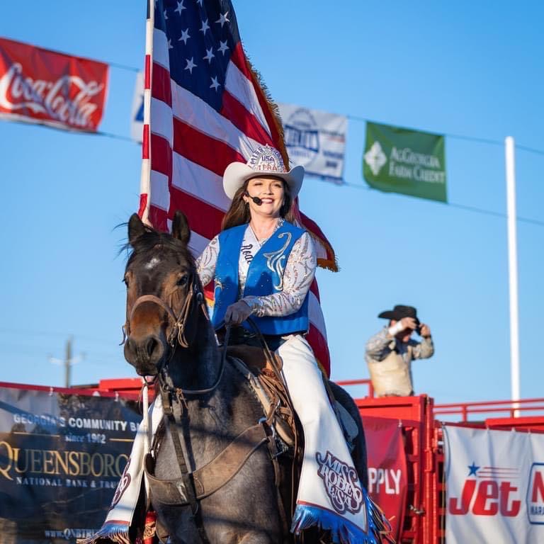 Woman in western attire riding a horse in front of an American flag, with a person taking photos in the background.