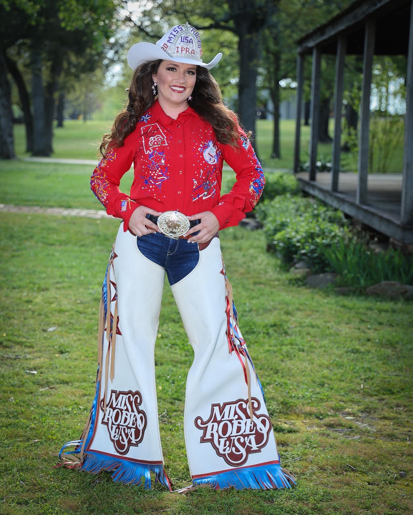 Young woman in cowboy hat and red shirt standing outdoors on grass, smiling, with trees and a building in background.