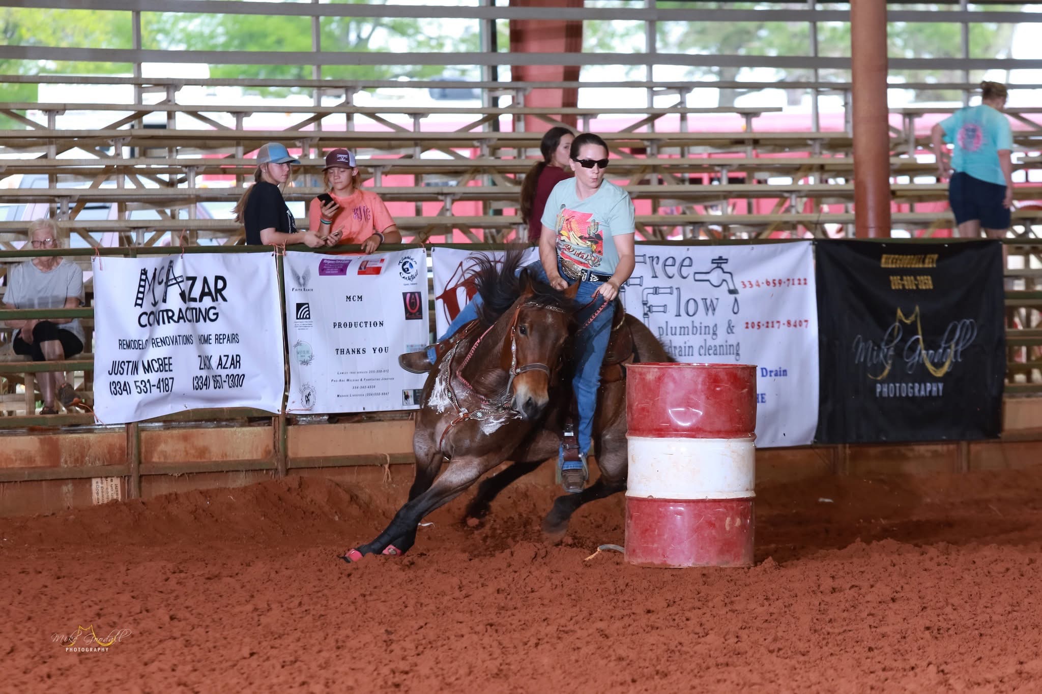 Person riding a bucking horse in an indoor arena with banners and spectators in the background.
