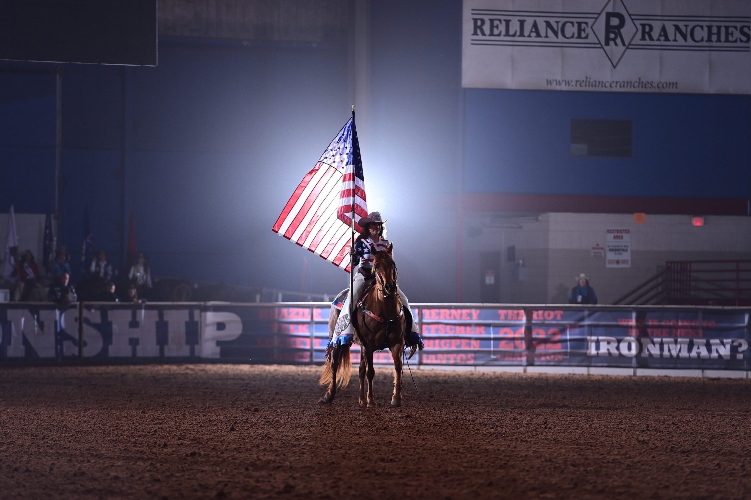 Person on horseback holding an American flag in an indoor arena with banners and audience in background.