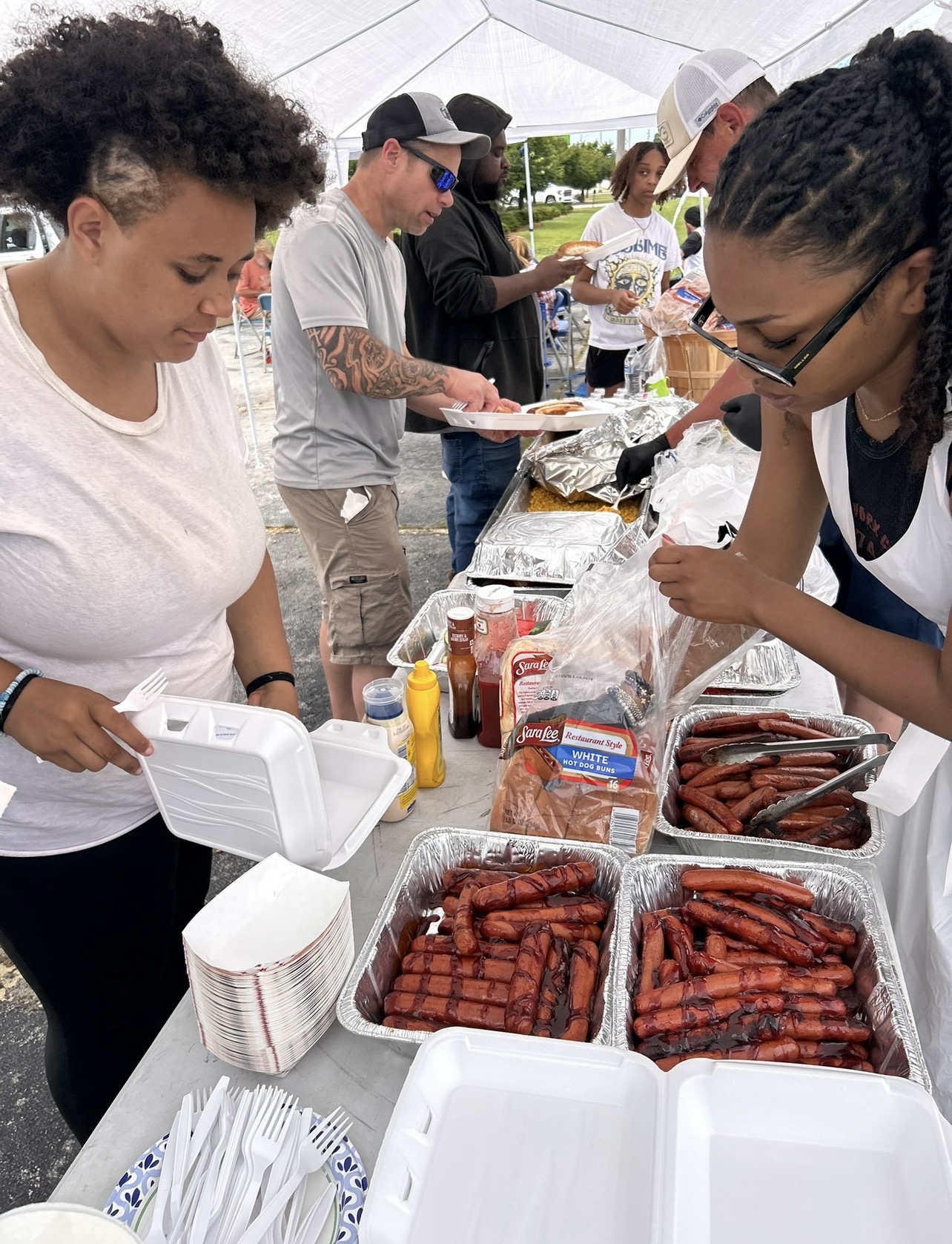 People gather around a table with food, preparing and serving grilled sausages outdoors under a tent.