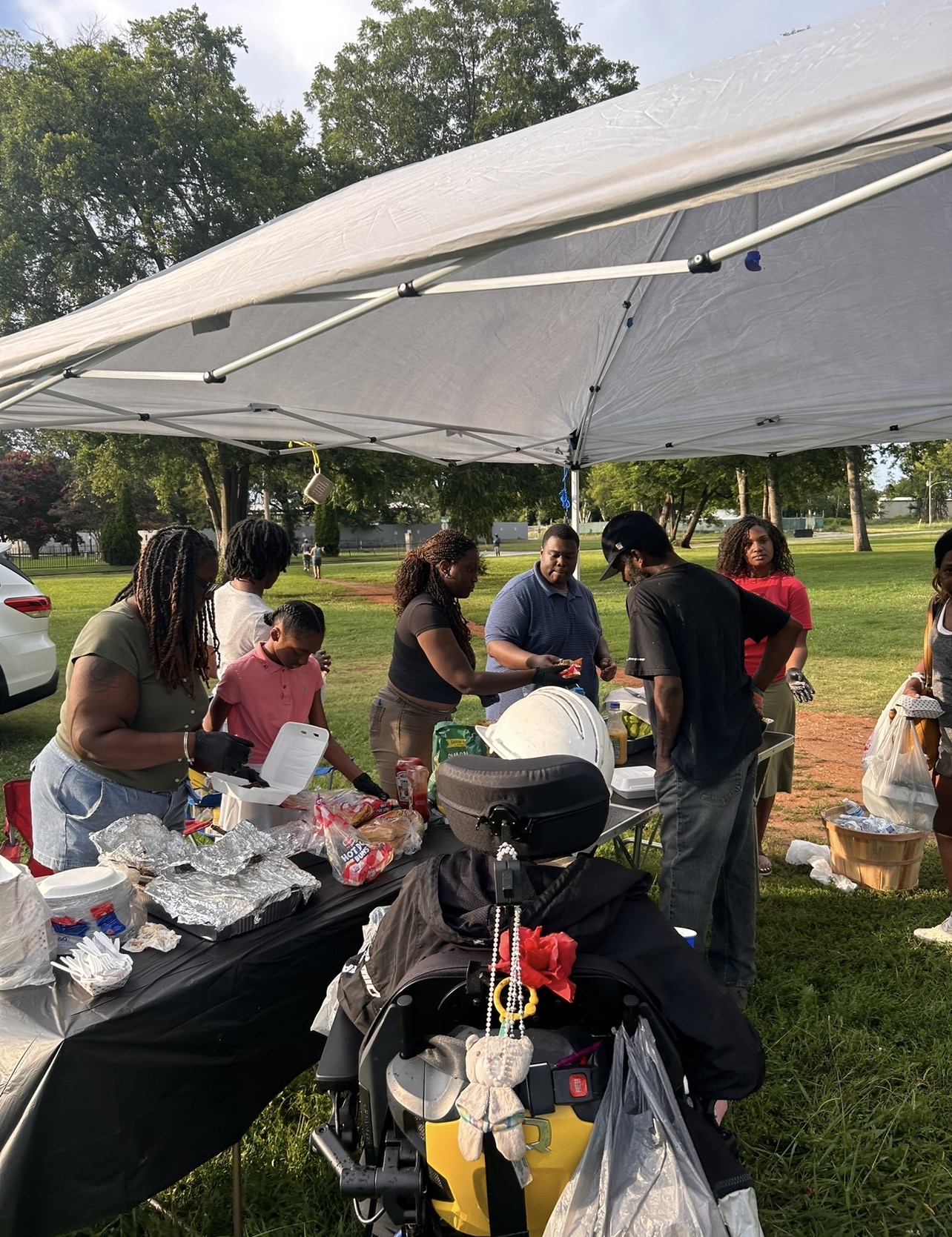 Group of people gathered under a white canopy in a park, with tables of food and supplies, trees in the background.