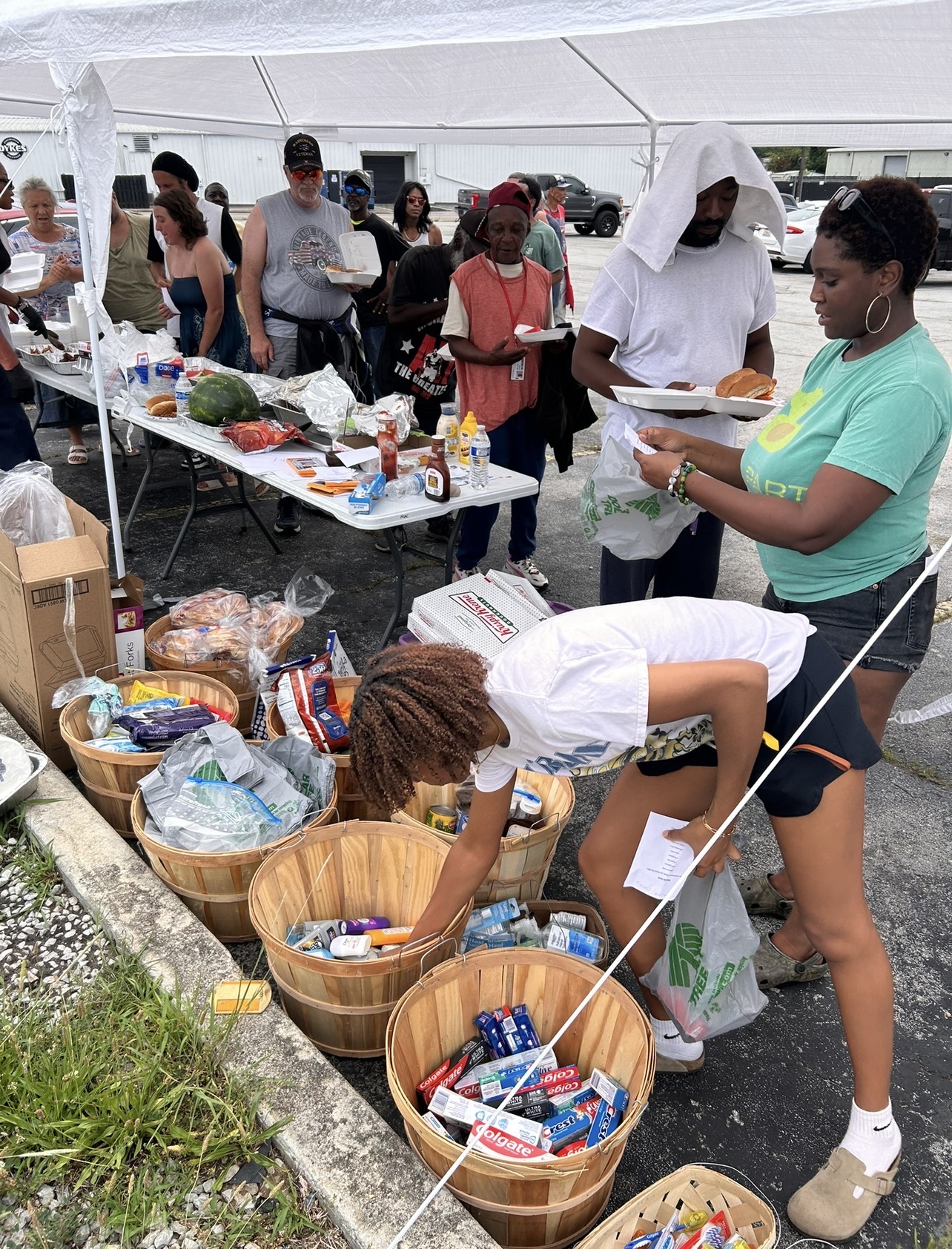 People gather at a food stall under a white tent, with baskets of snacks and drinks on a table.