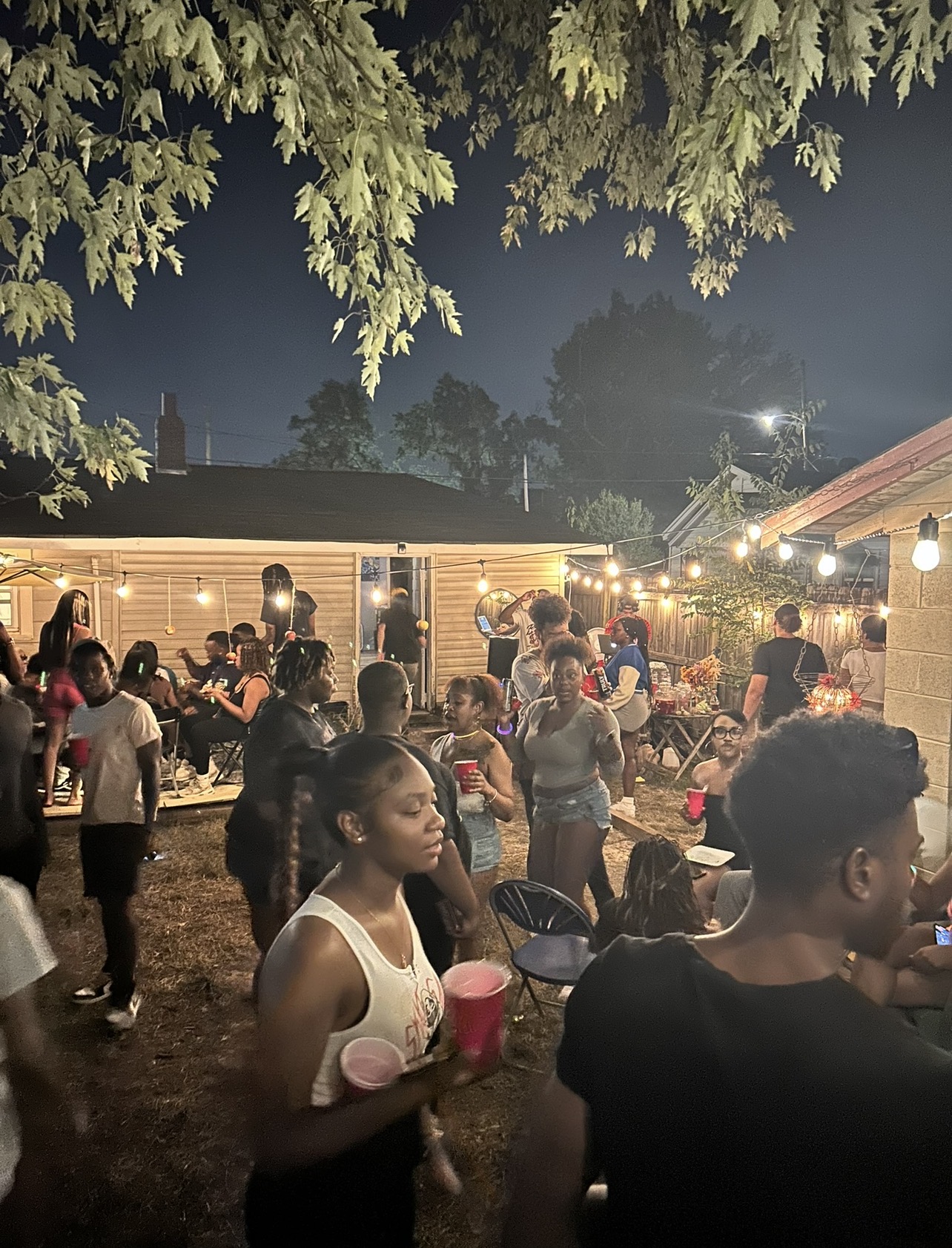 Outdoor gathering with people socializing and enjoying drinks at night, illuminated by string lights, with trees and a building in background.